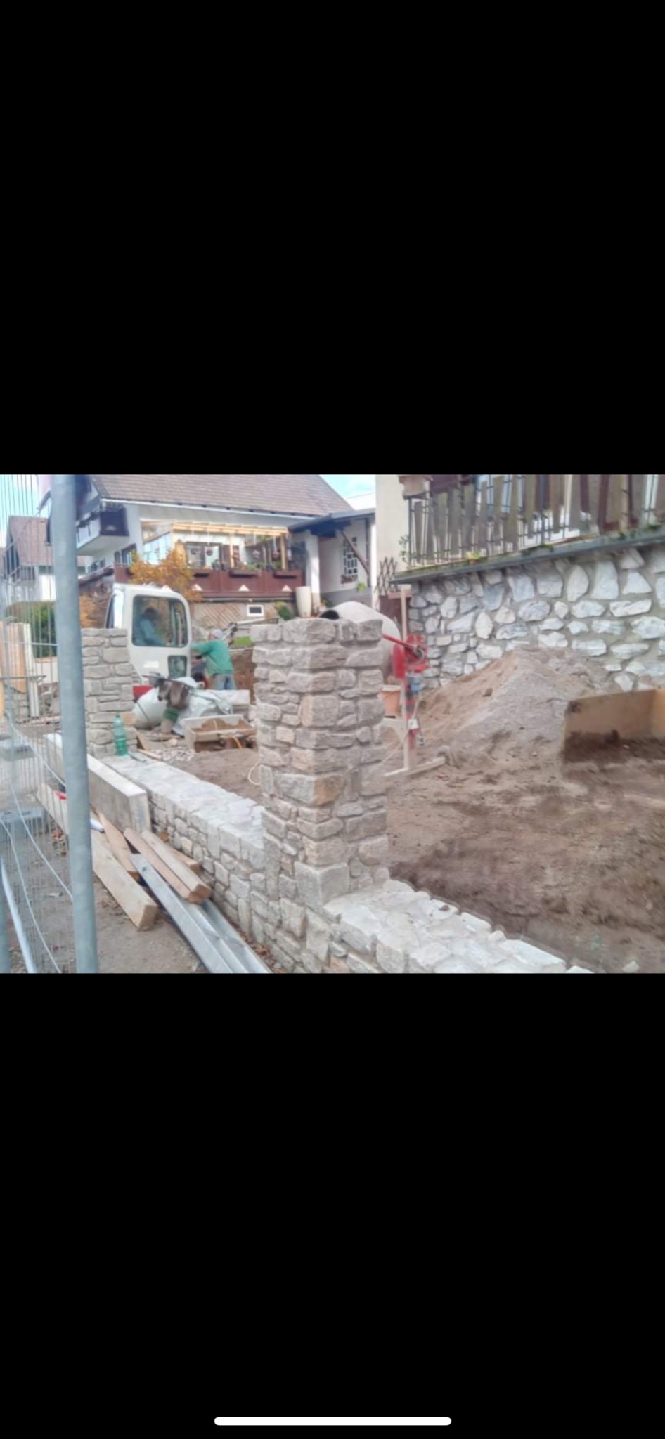 Budowa ogrodzenia z kamienia: widoczny fragment muru z kamiennymi słupkami, w tle pracownicy budowlani i betoniarka, na pierwszym planie deski i ziemia.