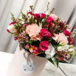 Ukffiecone Marta Sikorska - Kompozycja kwiatowa w pastelowym pudełku z logo kwiaciarni, zawierająca róże, goździki i alstromerie w odcieniach różu, bieli i kremu, na tle białej zasłony.