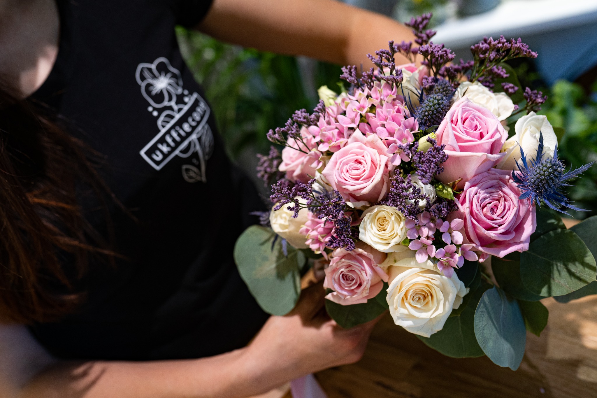 Bukiet ślubny z różowych i białych róż, fioletowych kwiatów i eukaliptusa trzymany w dłoniach na tle zieleni, wykonany przez kwiaciarnię z Katowic.