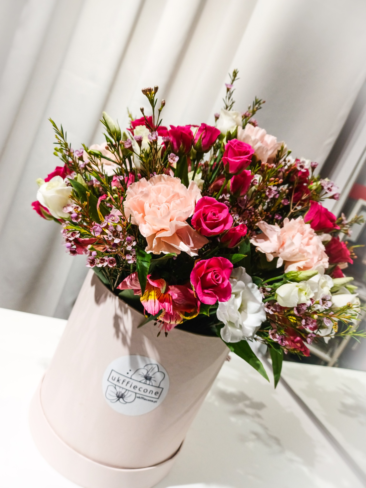 Kompozycja kwiatowa w pastelowym pudełku z logo kwiaciarni, zawierająca róże, goździki i alstromerie w odcieniach różu, bieli i kremu, na tle białej zasłony.