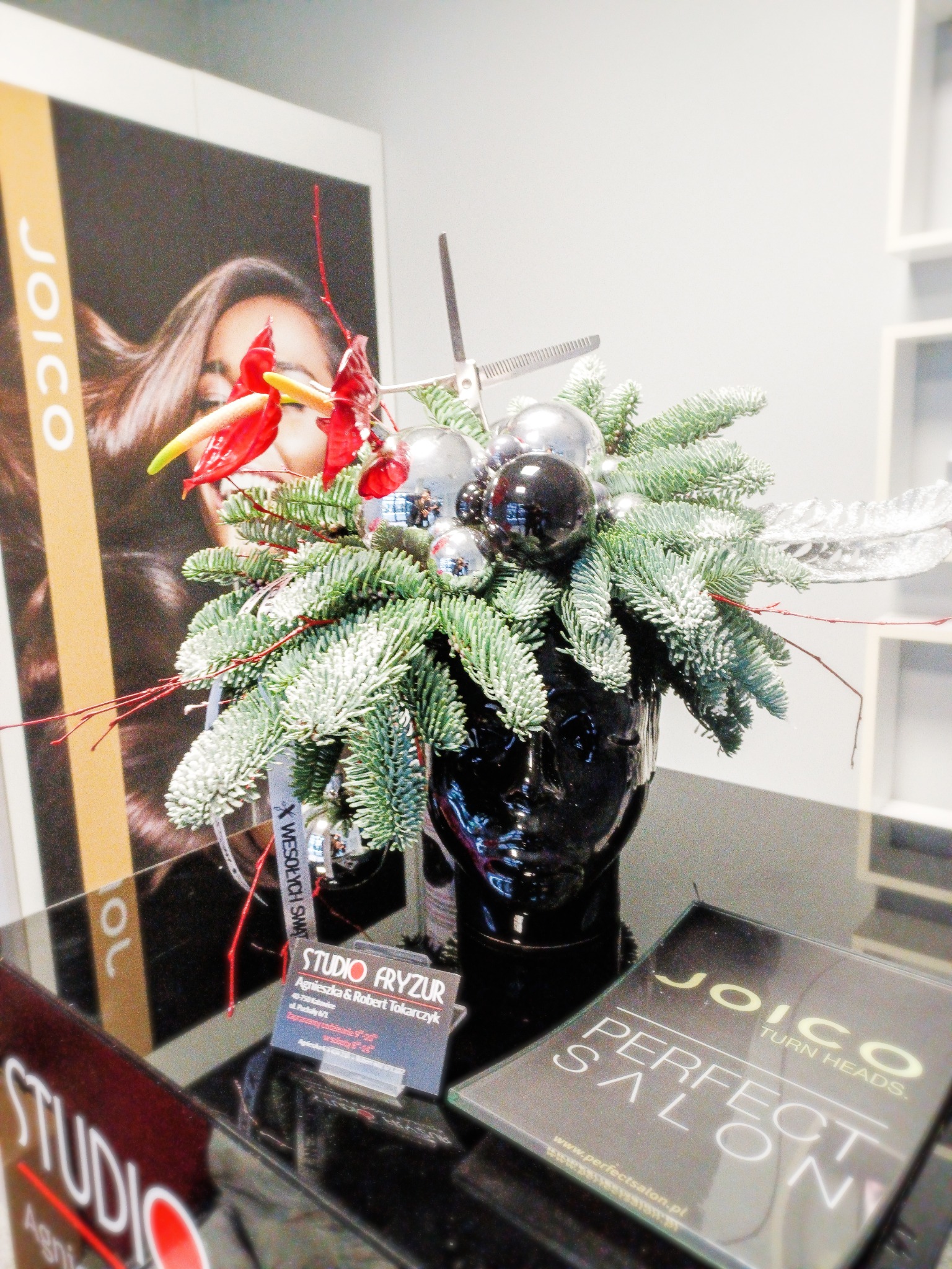 Dekoracja świąteczna w salonie fryzjerskim: stroik z gałązek, bombek i czerwonych anturiów w czarnej głowie-wazonie, obok reklama Joico i wizytówka salonu.