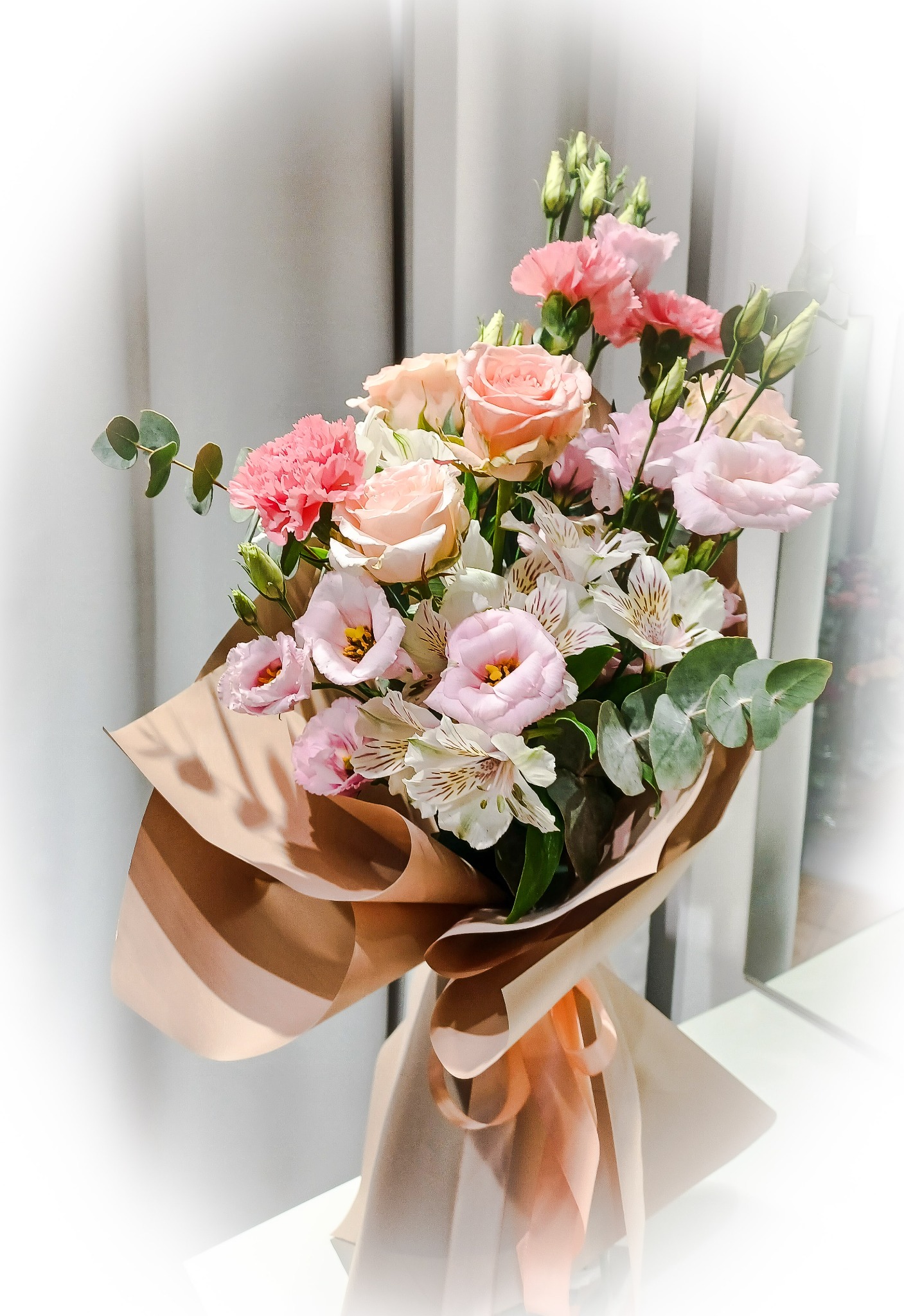 Delikatny bukiet z różowych róż, goździków i alstremerii, owinięty w beżowy papier i przewiązany wstążką, na tle białej zasłony.