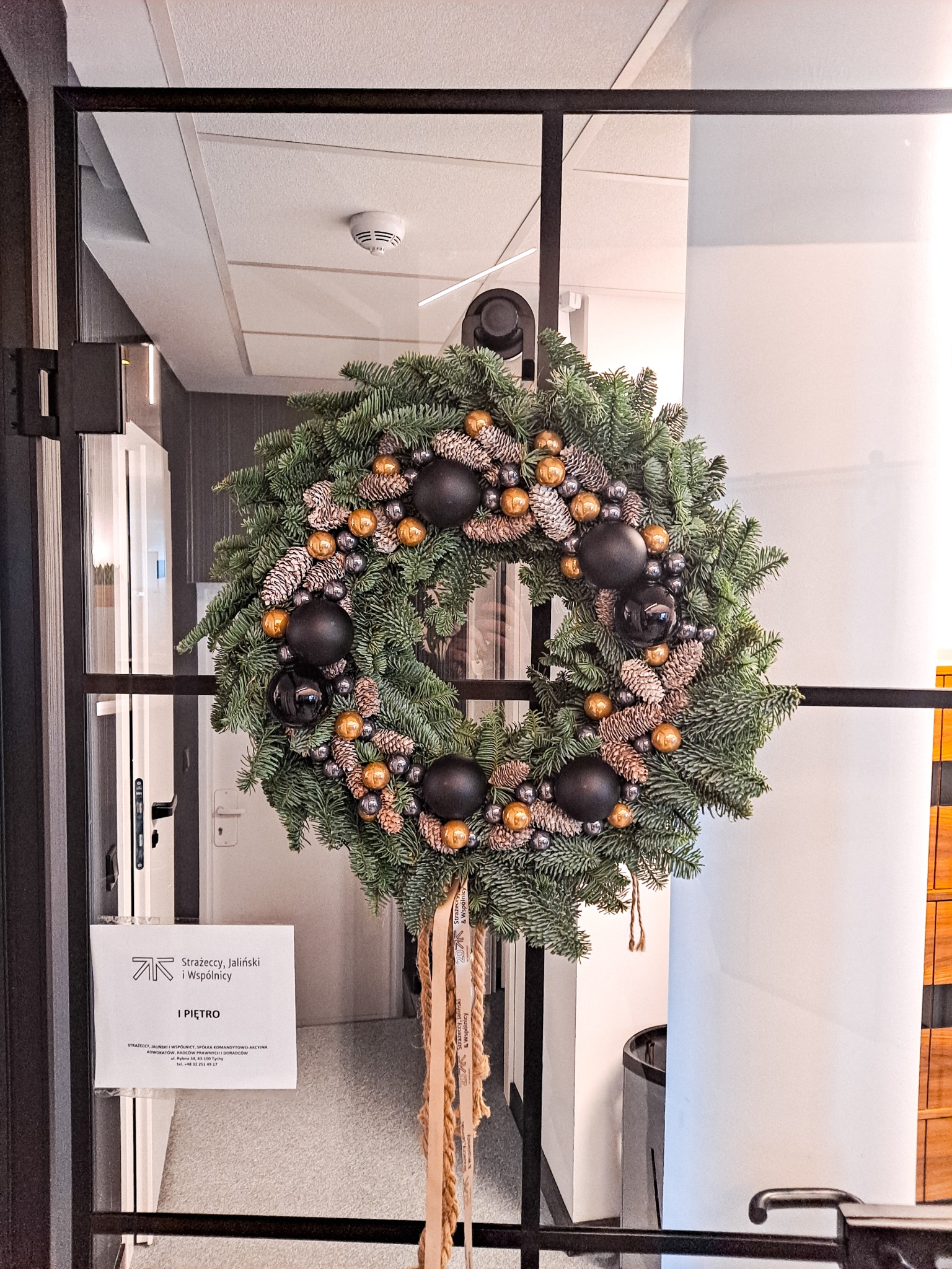 Świąteczny wieniec z ciemnozielonych gałązek, szyszek, złotych i czarnych bombek, zawieszony na szklanych drzwiach z czarną ramą w nowoczesnym biurze.