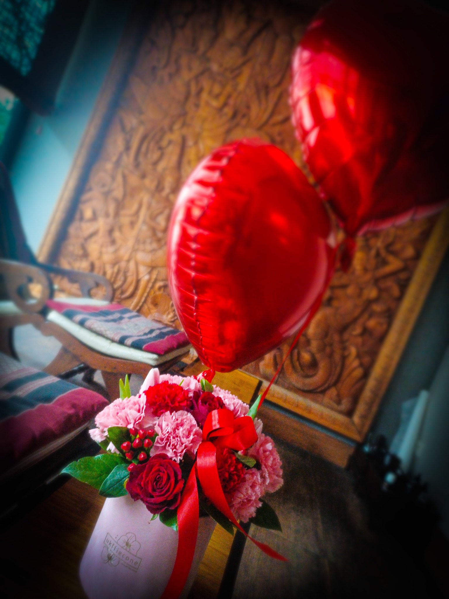 Kompozycja kwiatowa z czerwonymi różami i goździkami w jasnoróżowym pudełku, ozdobiona czerwoną wstążką, z dwoma czerwonymi balonami w tle i rzeźbioną drewnianą ramą na ścianie.