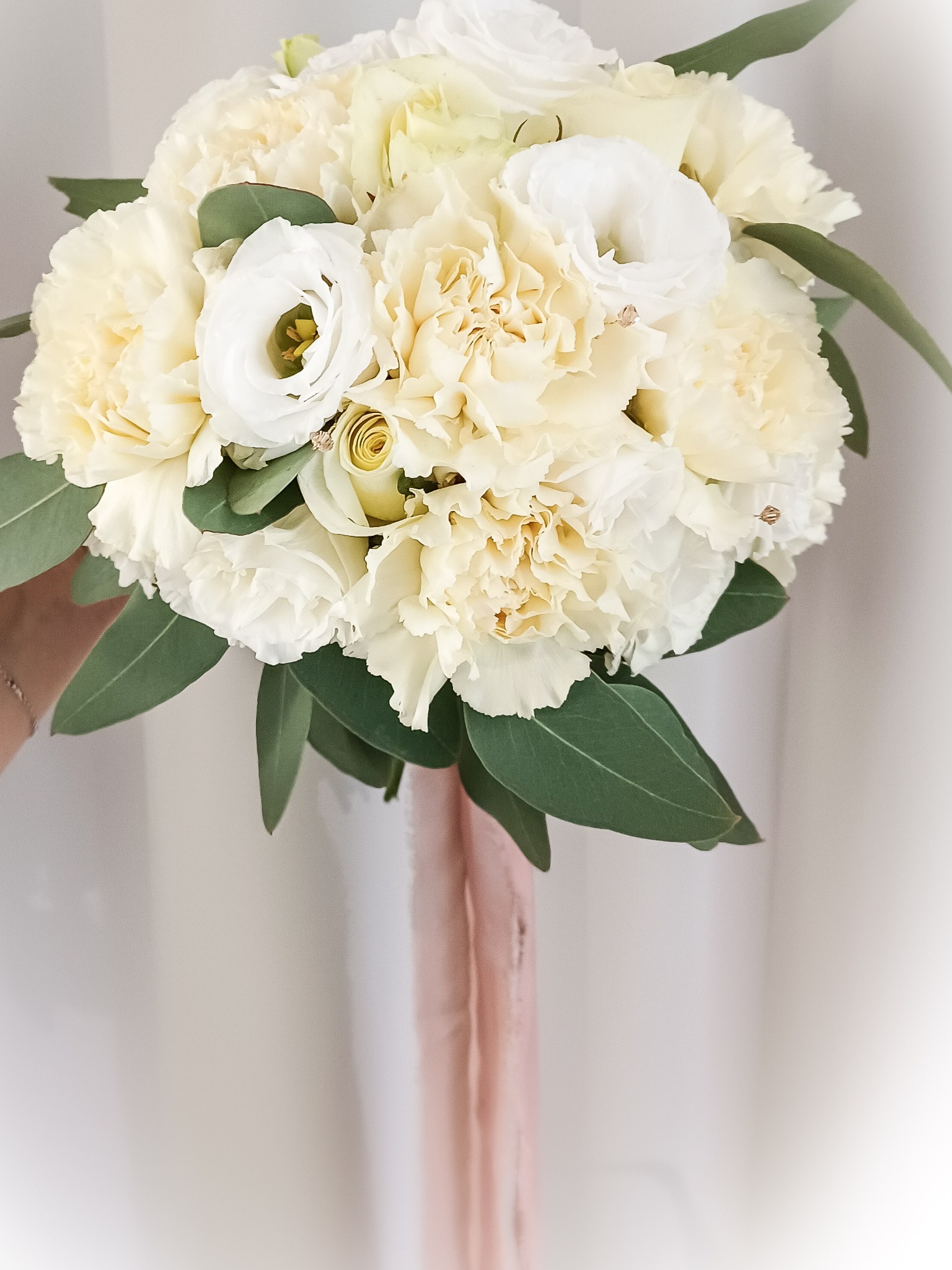 Delikatny bukiet ślubny z białych i kremowych goździków oraz eustomy, przewiązany różową wstążką, na jasnym tle.