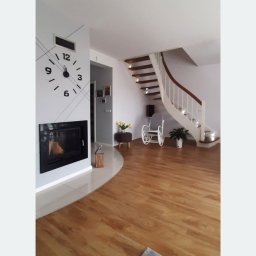 Tomasz Kalinowski - Jasne wnętrze domu z drewnianą podłogą, kominkiem z zegarem na ścianie i krętymi schodami. Widoczny fragment dywanu i dekoracje.