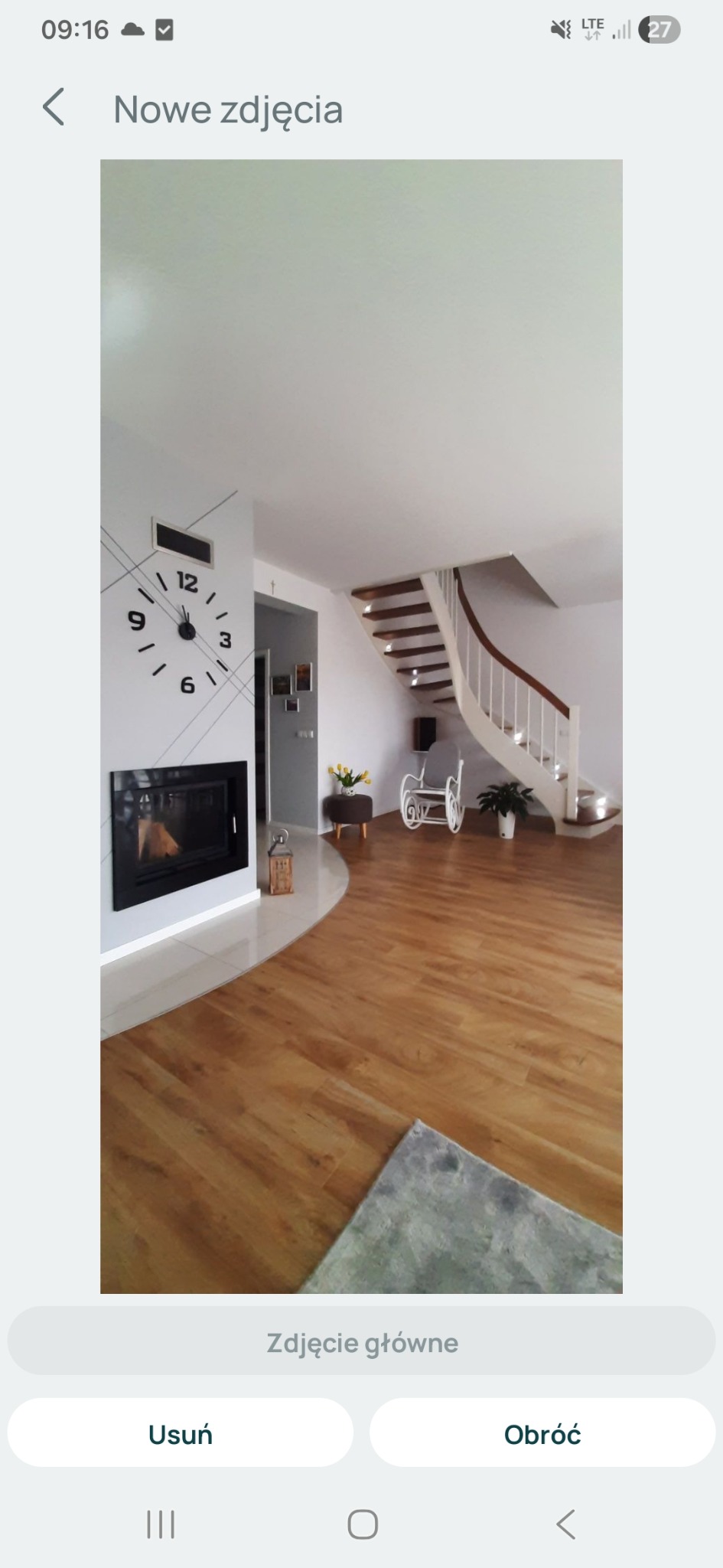 Jasne wnętrze domu z drewnianą podłogą, kominkiem z zegarem na ścianie i krętymi schodami. Widoczny fragment dywanu i dekoracje.