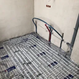 Białe rurki ogrzewania podłogowego ułożone na srebrnej folii z niebieską siatką w surowym pomieszczeniu z widocznymi instalacjami hydraulicznymi na ścianie.