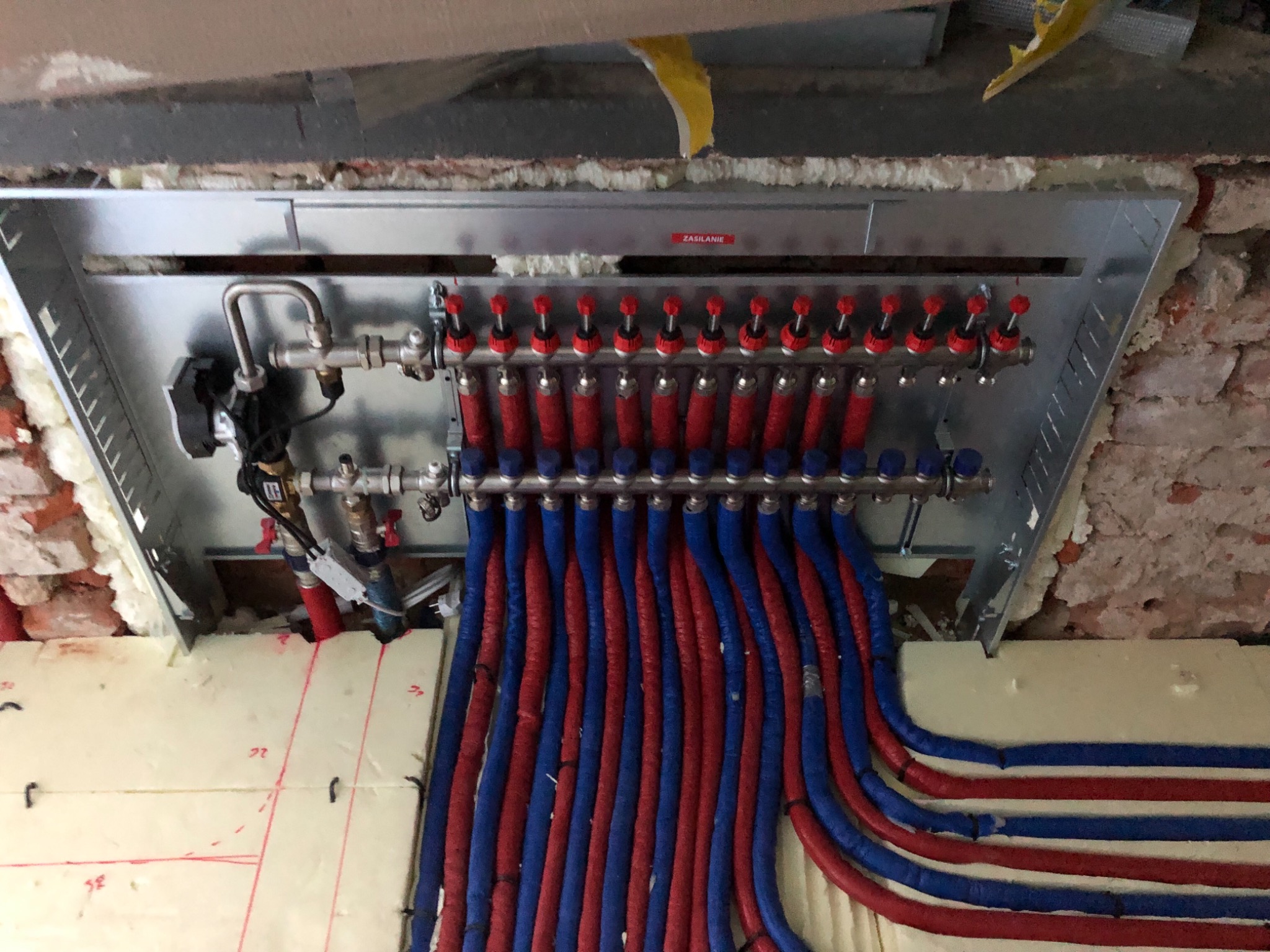 Rozdzielacz instalacji grzewczej podtynkowy z licznymi pętlami rur w kolorze czerwonym i niebieskim, zamontowany w ścianie z cegły, zabezpieczony pianką montażową.