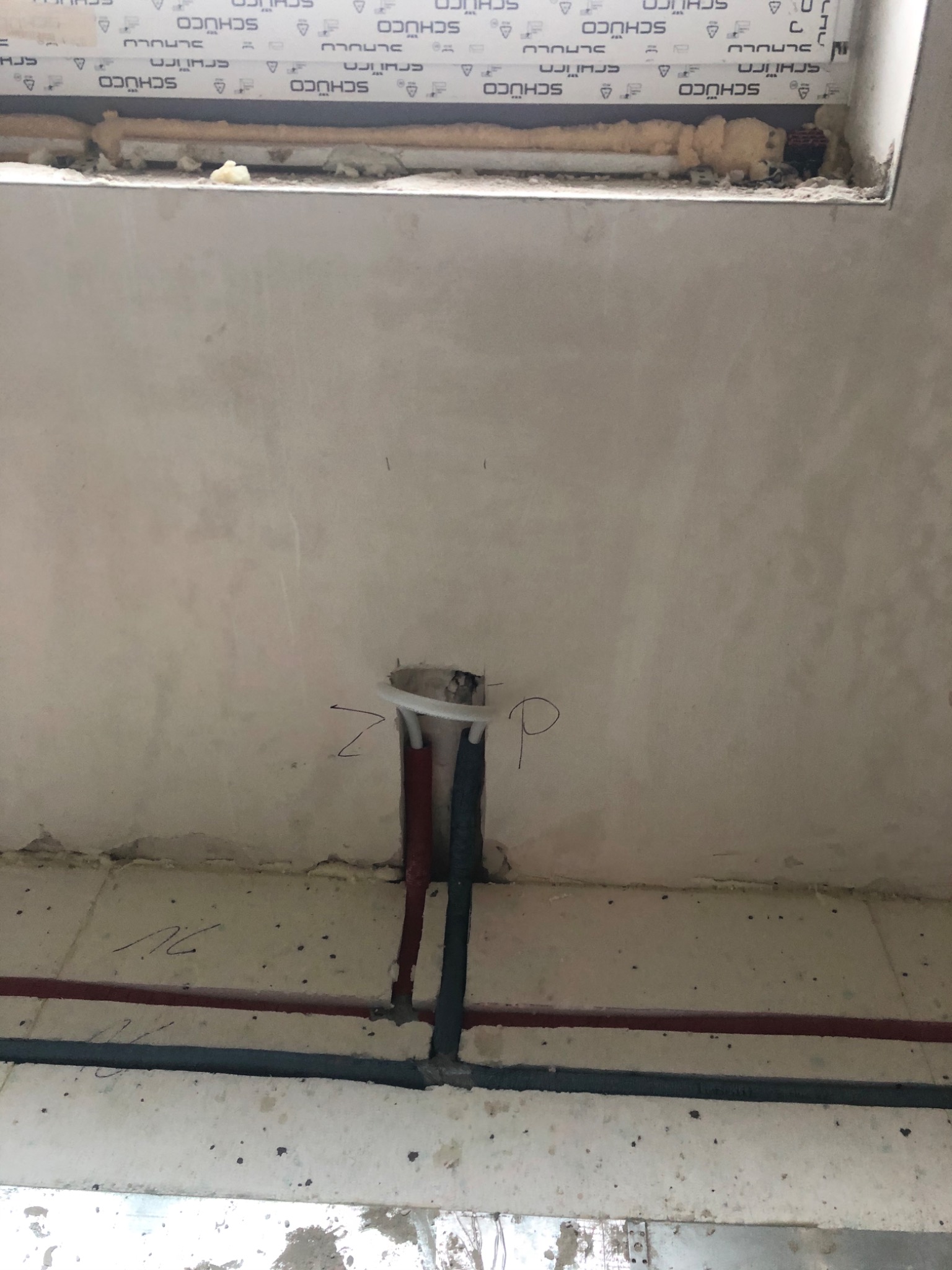 Surowy stan instalacji hydraulicznej w trakcie budowy: widoczne wystające rury, nieotynkowana ściana z napisem 'Z' i 'P', oraz fragment okna z pianką montażową i logo SCHÜCO.