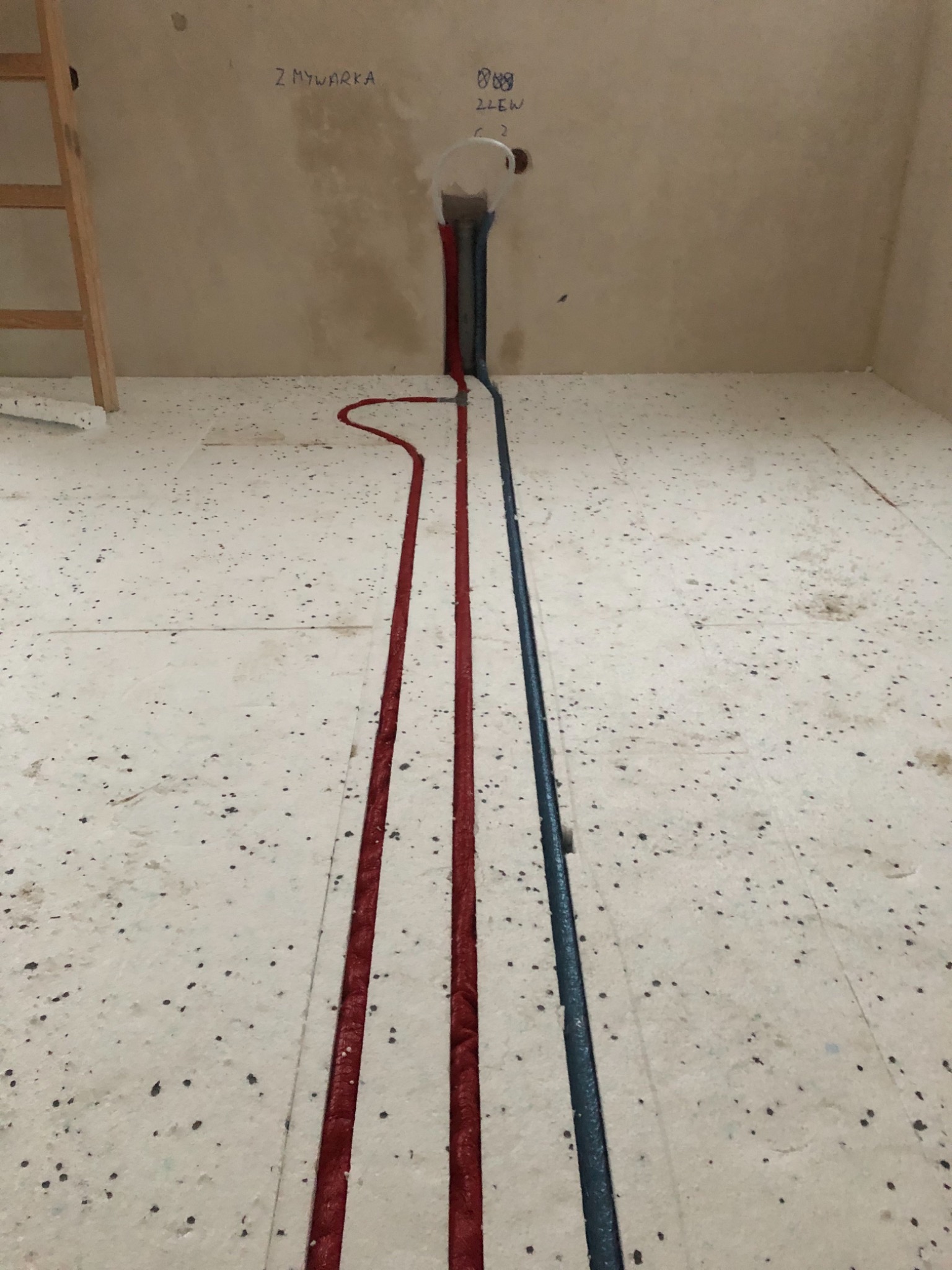 Podłogowe ogrzewanie podtynkowe w trakcie montażu, widoczne czerwone rury grzewcze i niebieska rura wodociągowa, ściana z napisem 'Z Mywarka'.