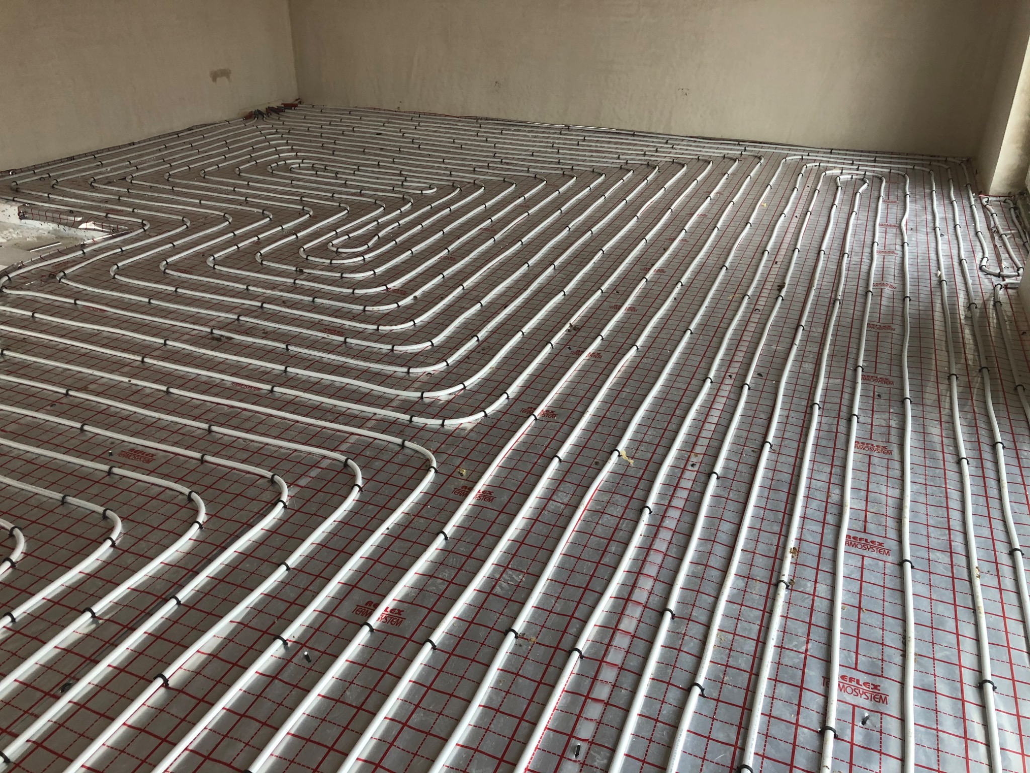 Białe rury ogrzewania podłogowego ułożone spiralnie na czerwonej siatce w surowym pomieszczeniu, widoczne czarne uchwyty mocujące rury.