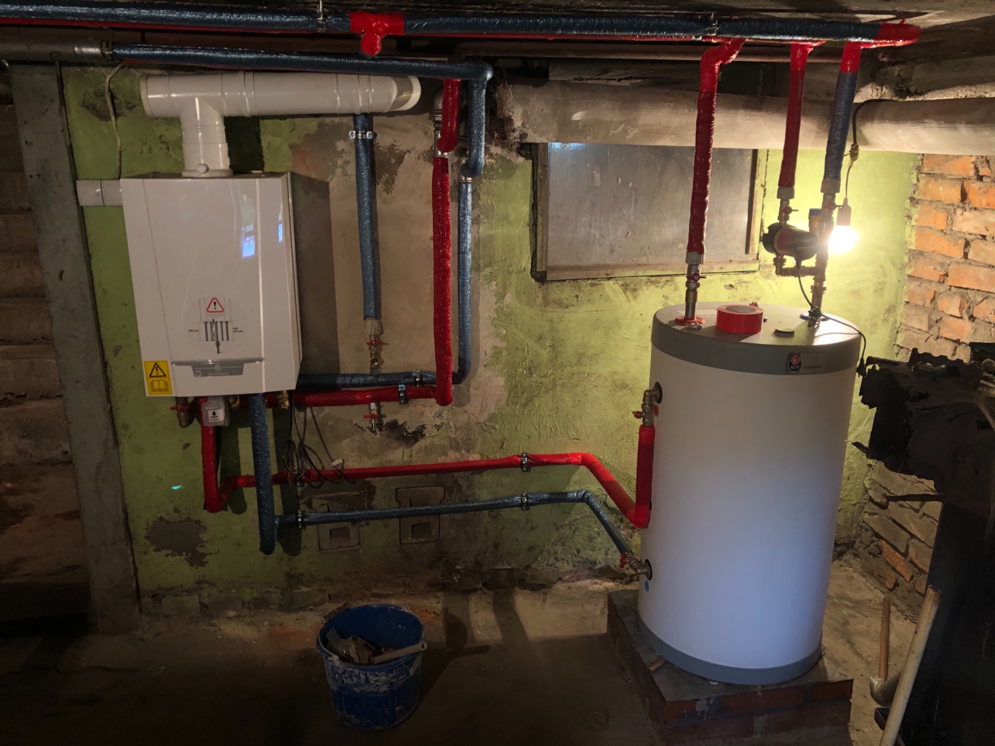 Biały piec gazowy wiszący na zielonej, nierównej ścianie piwnicy, z widocznymi rurami w izolacji czerwonej i niebieskiej, oraz stojący obok biały zbiornik na wodę.
