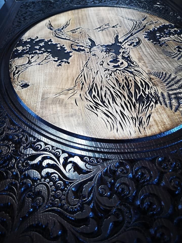 Drewniane drzwi z ręcznie rzeźbionym motywem jelenia na tle lasu i gór w okrągłej ramie z ozdobnymi ornamentami.