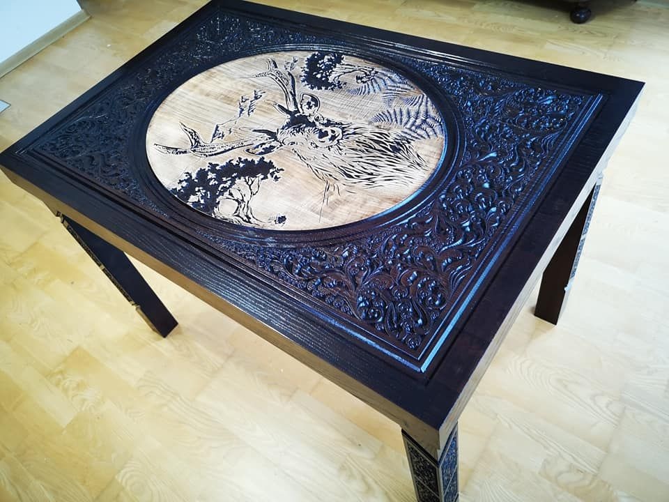 Kwadratowy stolik kawowy z ciemnego drewna z rzeźbionym wzorem florystycznym na obrzeżach i okrągłym blatem z jasnego drewna z wygrawerowanym motywem jelenia w lesie.