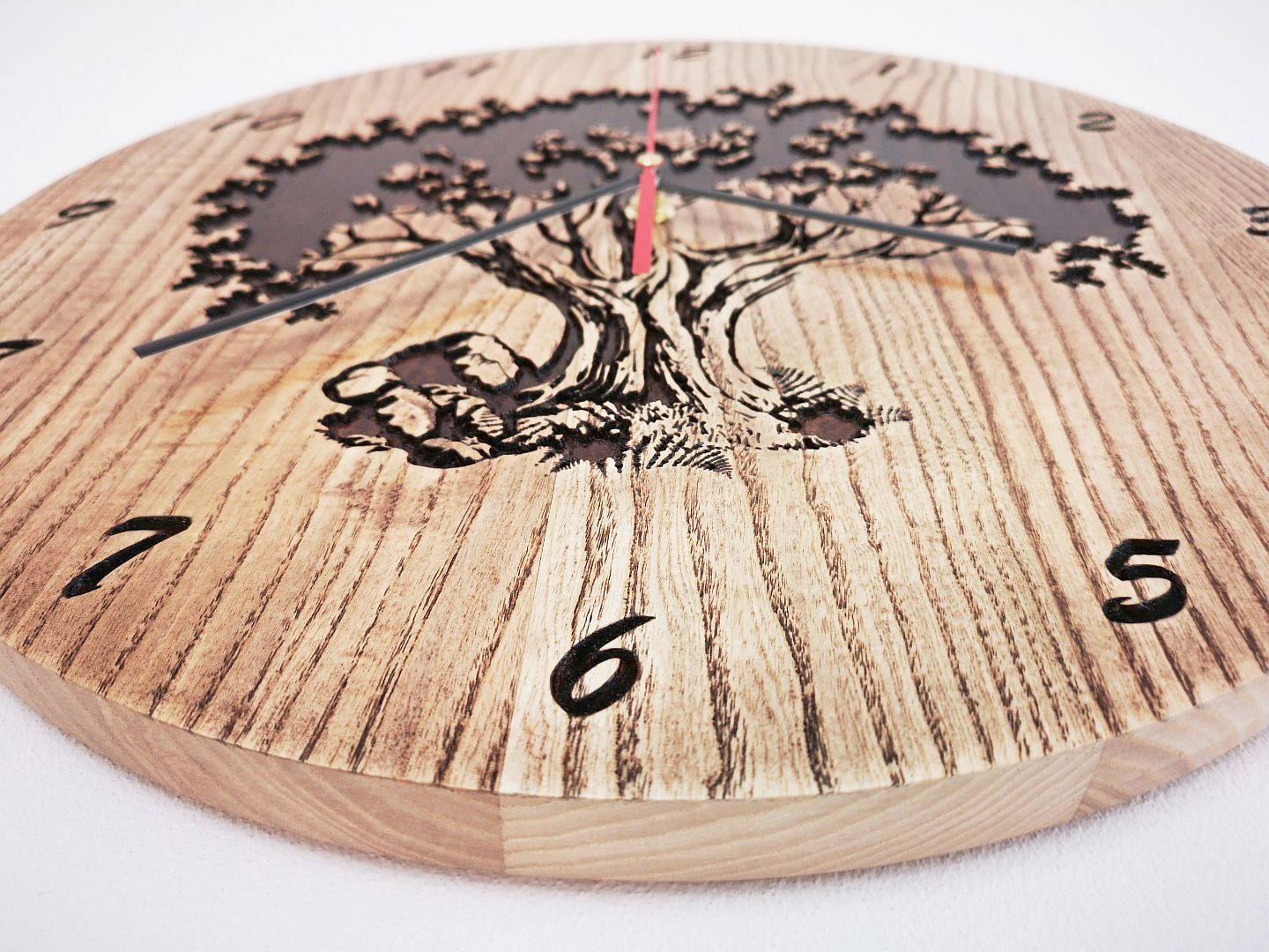 Okrągły zegar z jasnego drewna z wygrawerowanym motywem drzewa i cyframi, wskazówki czarne i czerwona, widoczne słoje drewna.