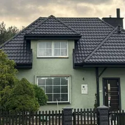Dom z nowym, grafitowym dachem z blachodachówki GERMAN 9005 w Ulanowie. Metalowe rynny BRYZA. Widok z przodu, zieleń w tle.