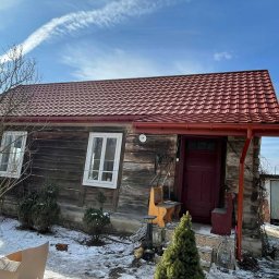 SMARTDACH Sp. z o.o. - Ceglasty blachodach na drewnianym domu z oknami i rynnami w tym samym kolorze. Wokół resztki śniegu i roślinność. Błękitne niebo z smugą kondensacyjną.