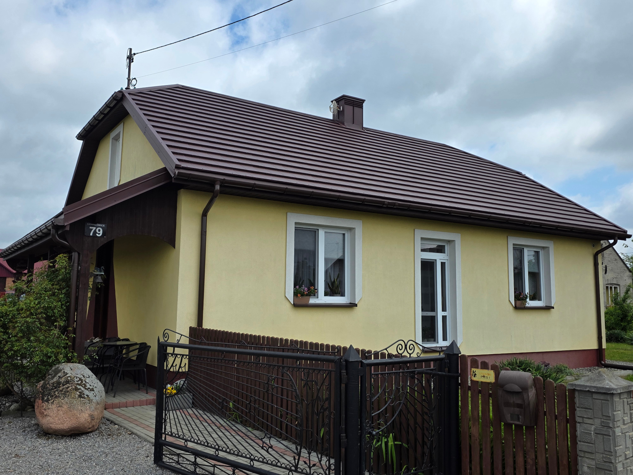 Żółty dom jednorodzinny z brązowym dachem z blachodachówki płaskiej, brązowe rynny, biała stolarka okienna, ozdobne skrzynki z kwiatami pod oknami.