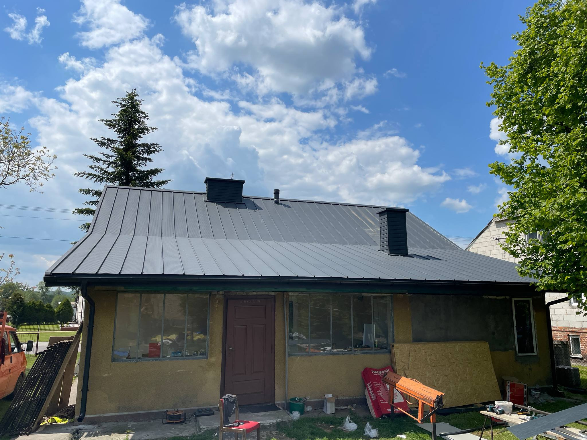 Dom z nowym, szarym dachem z blachy trapezowej, widoczne kominy. W tle błękitne niebo z chmurami i drzewa. Częściowo widoczne materiały budowlane.