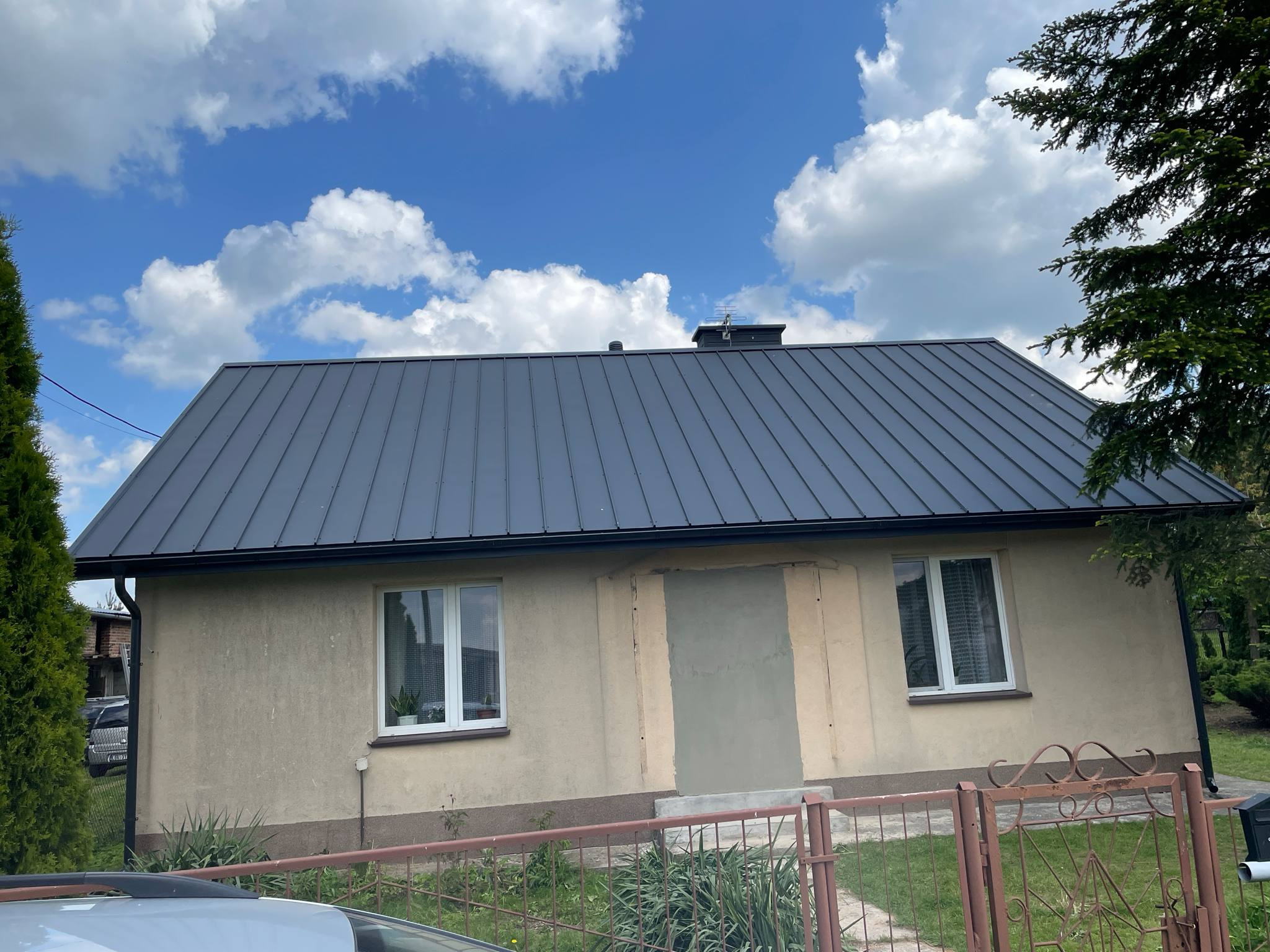 Dom z nowym, grafitowym dachem z blachy na rąbek stojący, model TP-26 w ral 7016 ultramat, w słoneczny dzień. Widok na fragment ogrodzenia i auta.