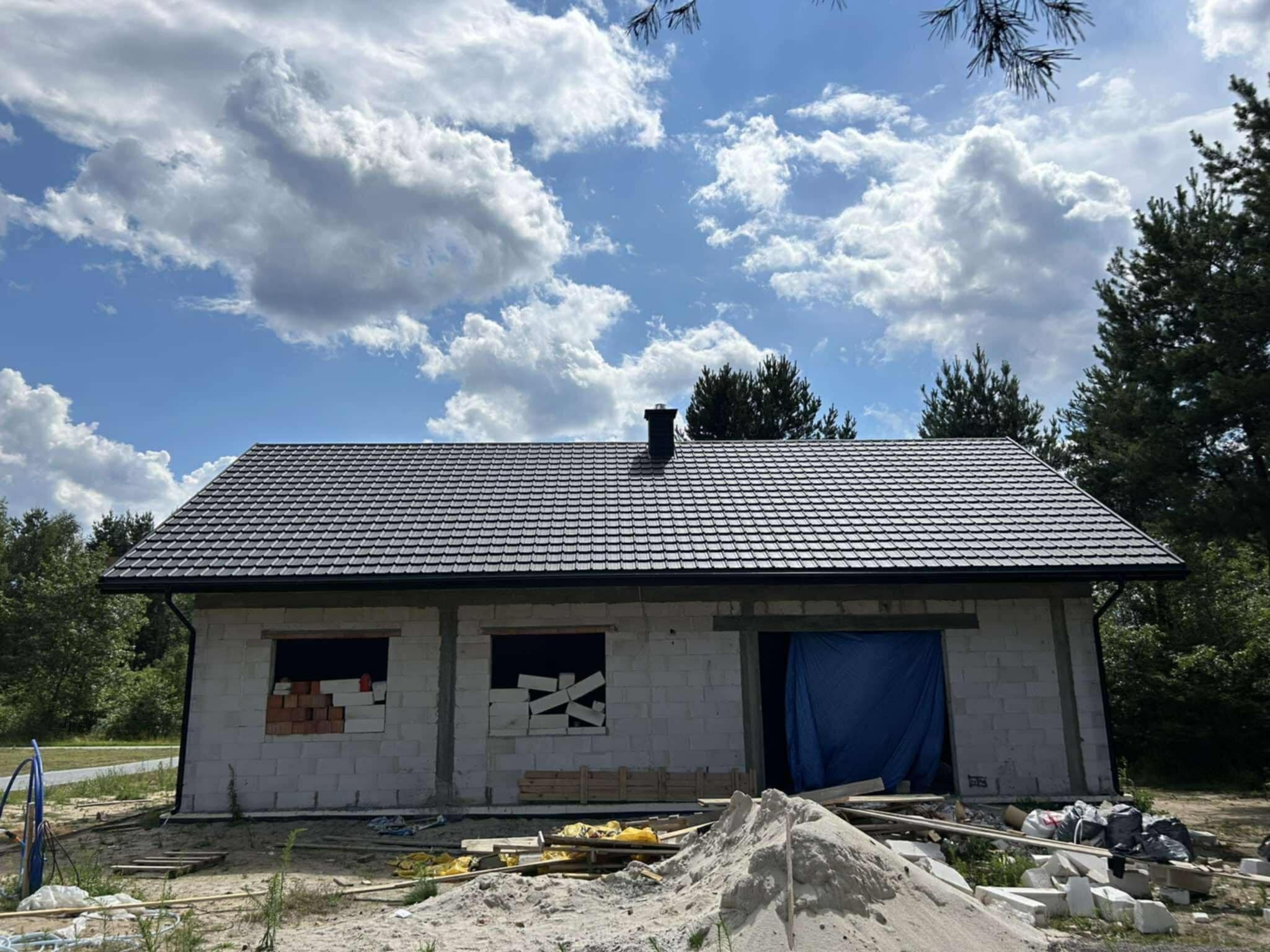 Czarny dach z blachodachówki, model FINN 2.0, na surowym budynku z białego bloczka. Częściowo zamurowane okna. W tle błękitne niebo z białymi chmurami.