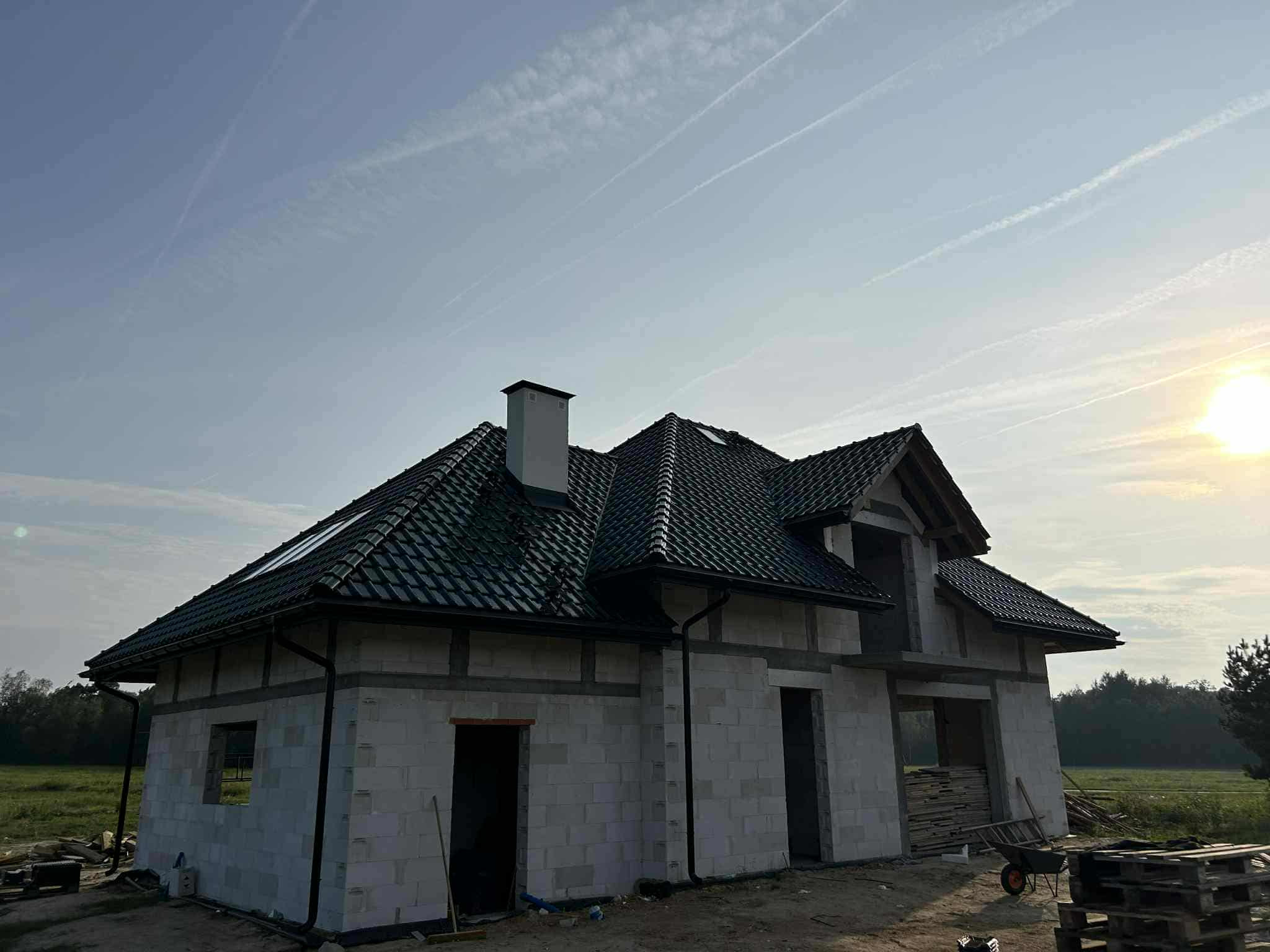 Dom w trakcie budowy z czarną, błyszczącą dachówką ceramiczną Braas Rubin 13V. Widoczne niezadaszone otwory okienne i drzwiowe, tło stanowi łąka i niebo.