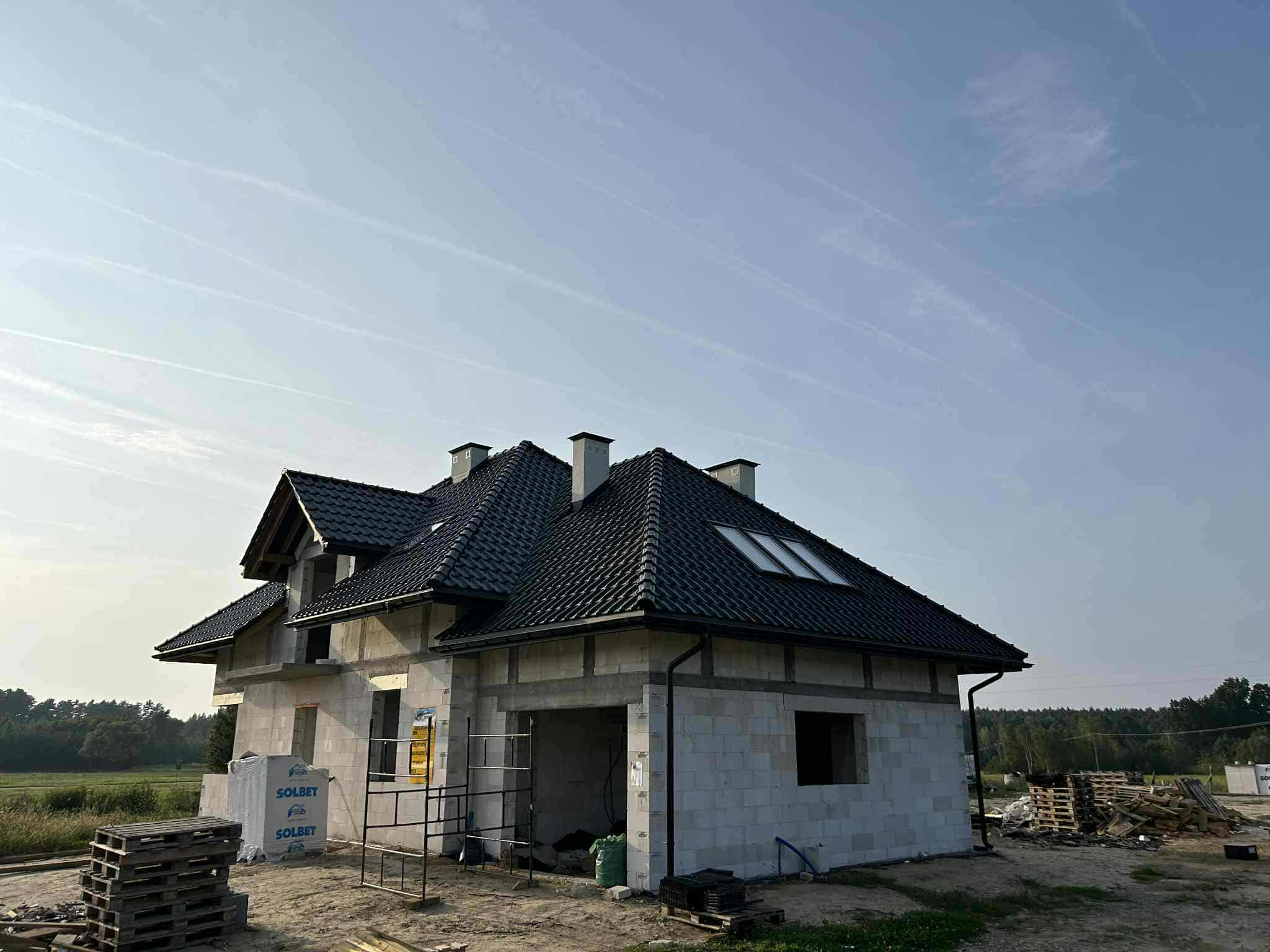 Dom w budowie z czarną, błyszczącą dachówką ceramiczną Braas Rubin 13V. Widoczny fragment elewacji i okna dachowe. W tle błękitne niebo i zieleń.