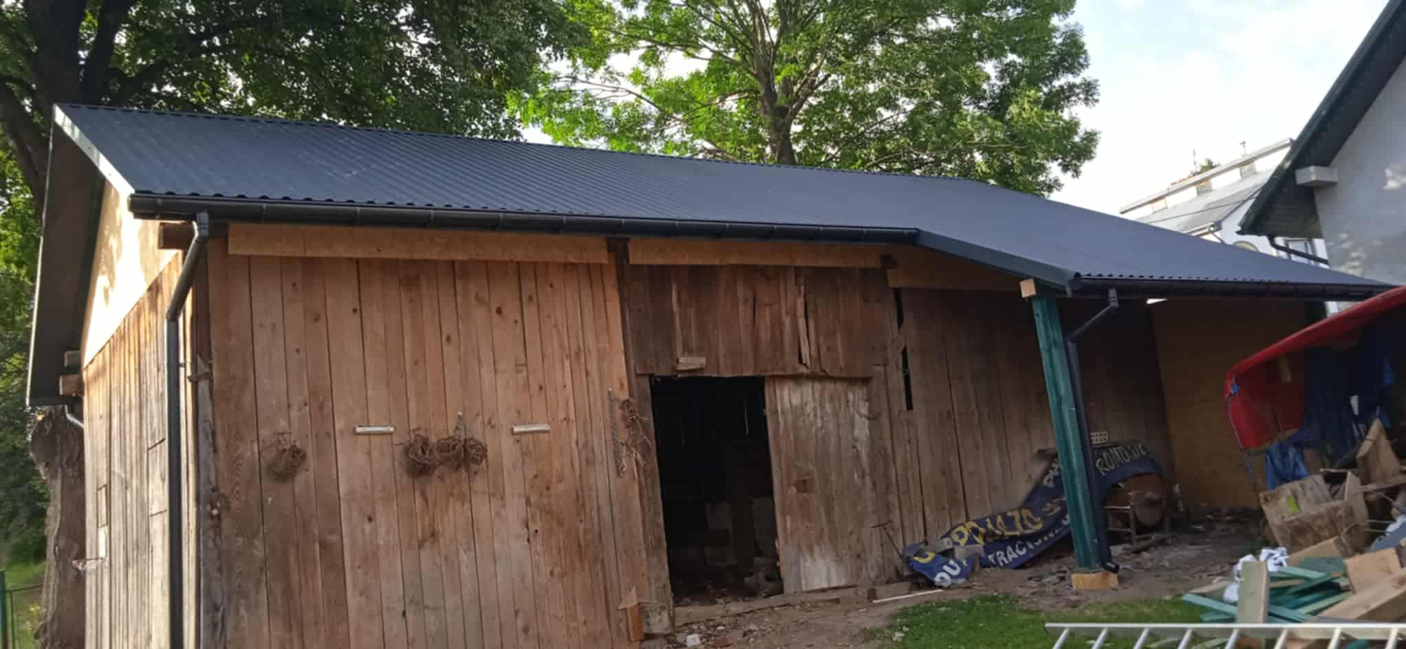 Stodoła po remoncie z nowym dachem z blachy trapezowej w kolorze ciemnoszarym. Widoczne rynny i drewniana konstrukcja budynku. Tło: drzewa.