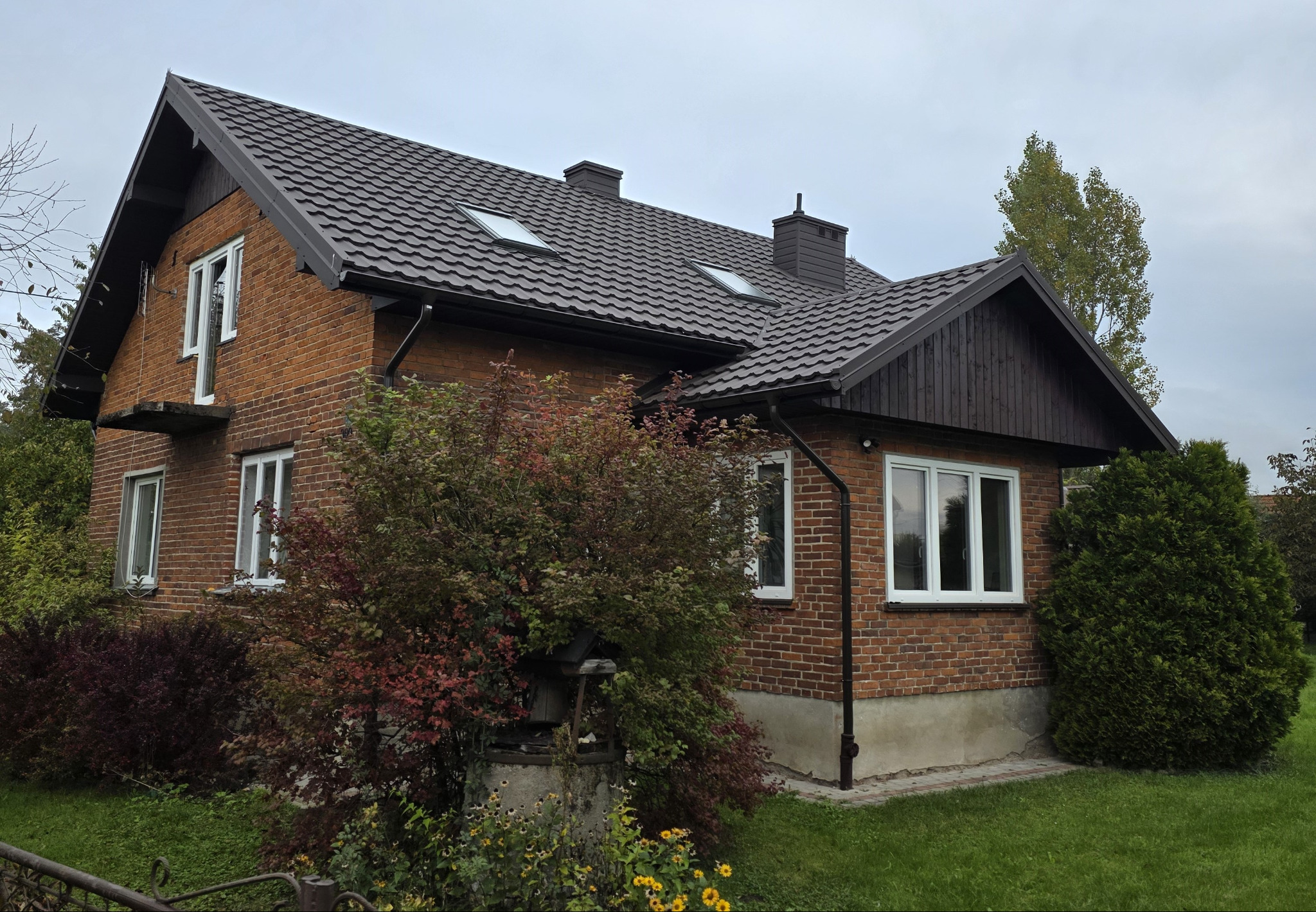 Dom z nowym, ciemnobrązowym dachem z blachodachówki Karpatia i metalowymi rynnami Bryza. Widoczne okna dachowe Fakro i ceglana elewacja budynku.