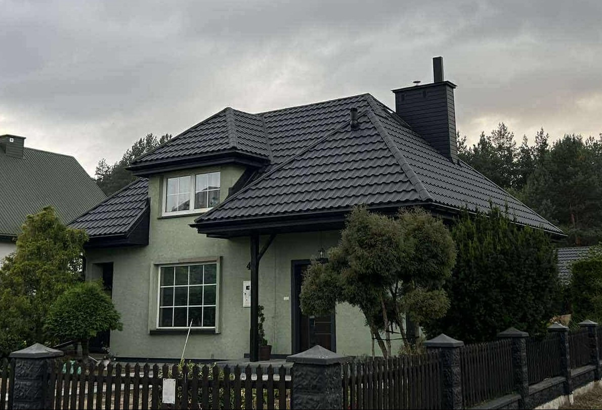 Dom z nowym, ciemnoszarym dachem z blachodachówki GERMAN 9005 PladurWrinkle mat, rynnami BRYZA i kominem w tym samym kolorze. Widok zza drewnianego płotu.
