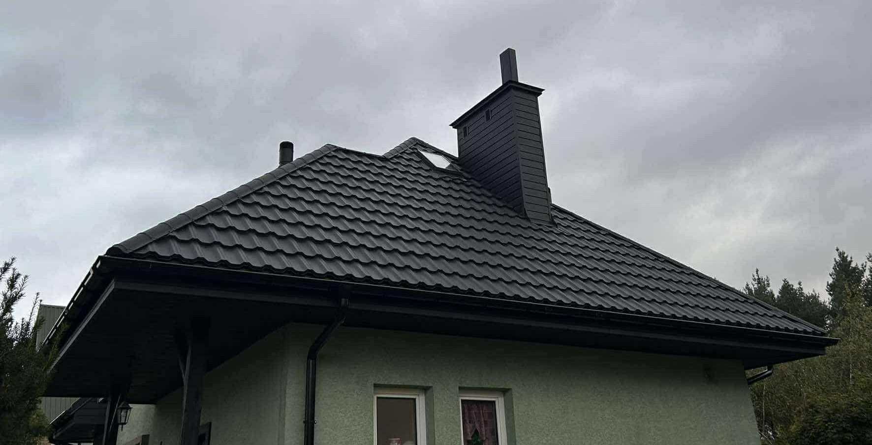 Czarny dach z blachodachówki GERMAN 9005 PladurWrinkle mat, z metalowymi rynnami BRYZA, na tle pochmurnego nieba i fragmentu elewacji budynku z dwoma oknami.