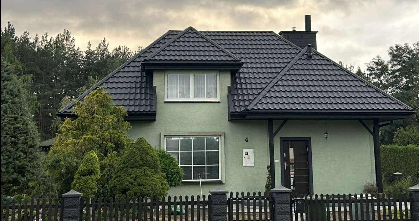 Dom z nowym, grafitowym dachem z blachodachówki GERMAN 9005 w Ulanowie. Metalowe rynny BRYZA. Widok z przodu, zieleń w tle.