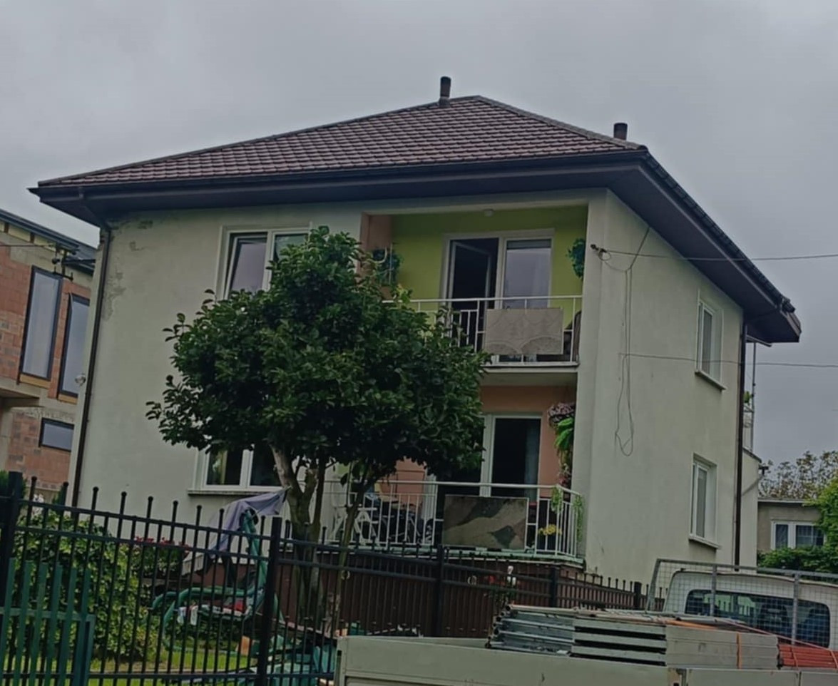 Dwupiętrowy dom z nowym, brązowym dachem z blachodachówki i metalowymi rynnami, widoczny zza ogrodzenia i drzewa. Częściowo widoczny samochód dostawczy.