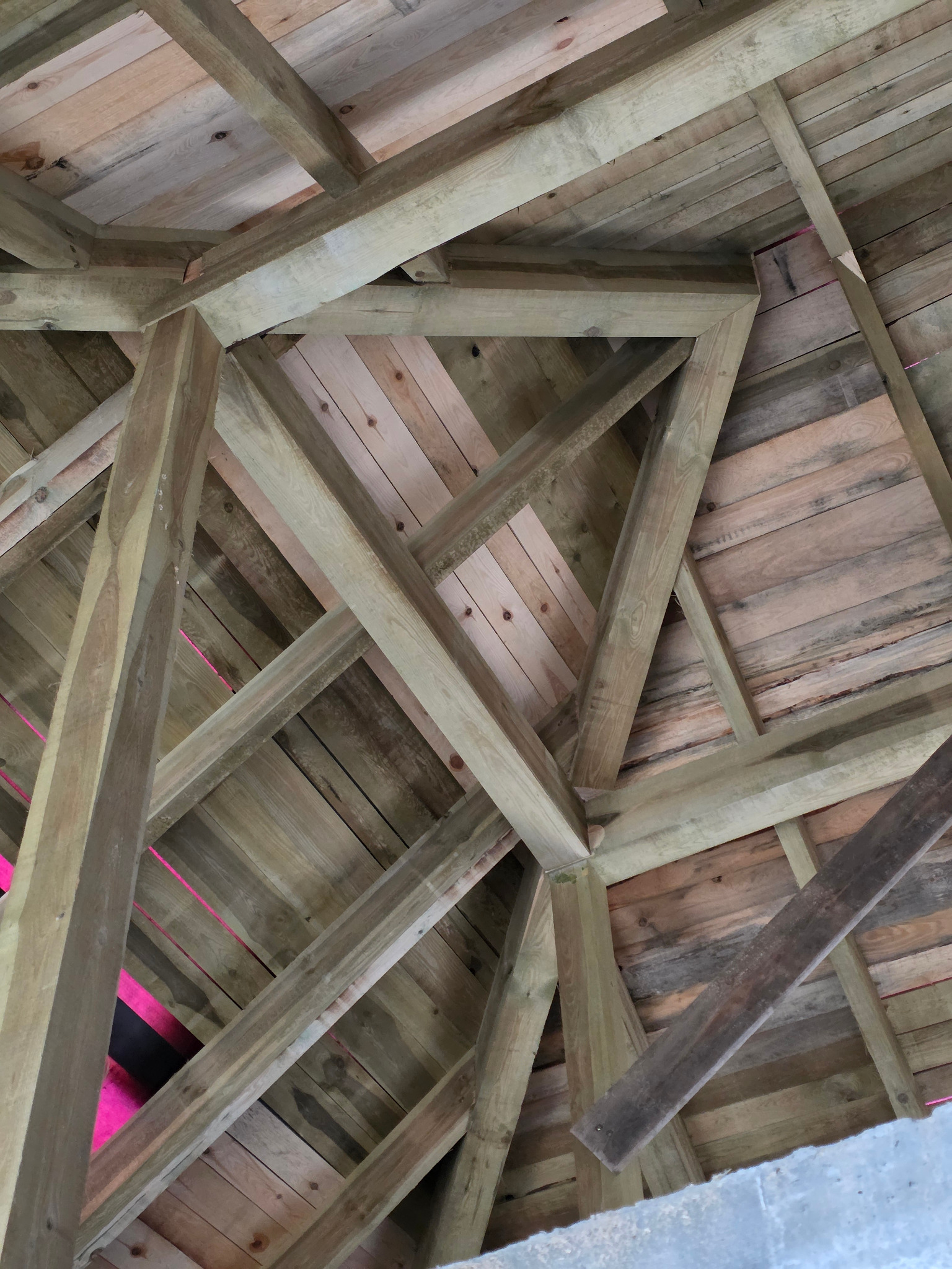 Wnętrze konstrukcji dachu z drewnianą więźbą i deskowaniem, widok z dołu, ukazujący skomplikowany układ belek i desek. Widoczne elementy mocujące.