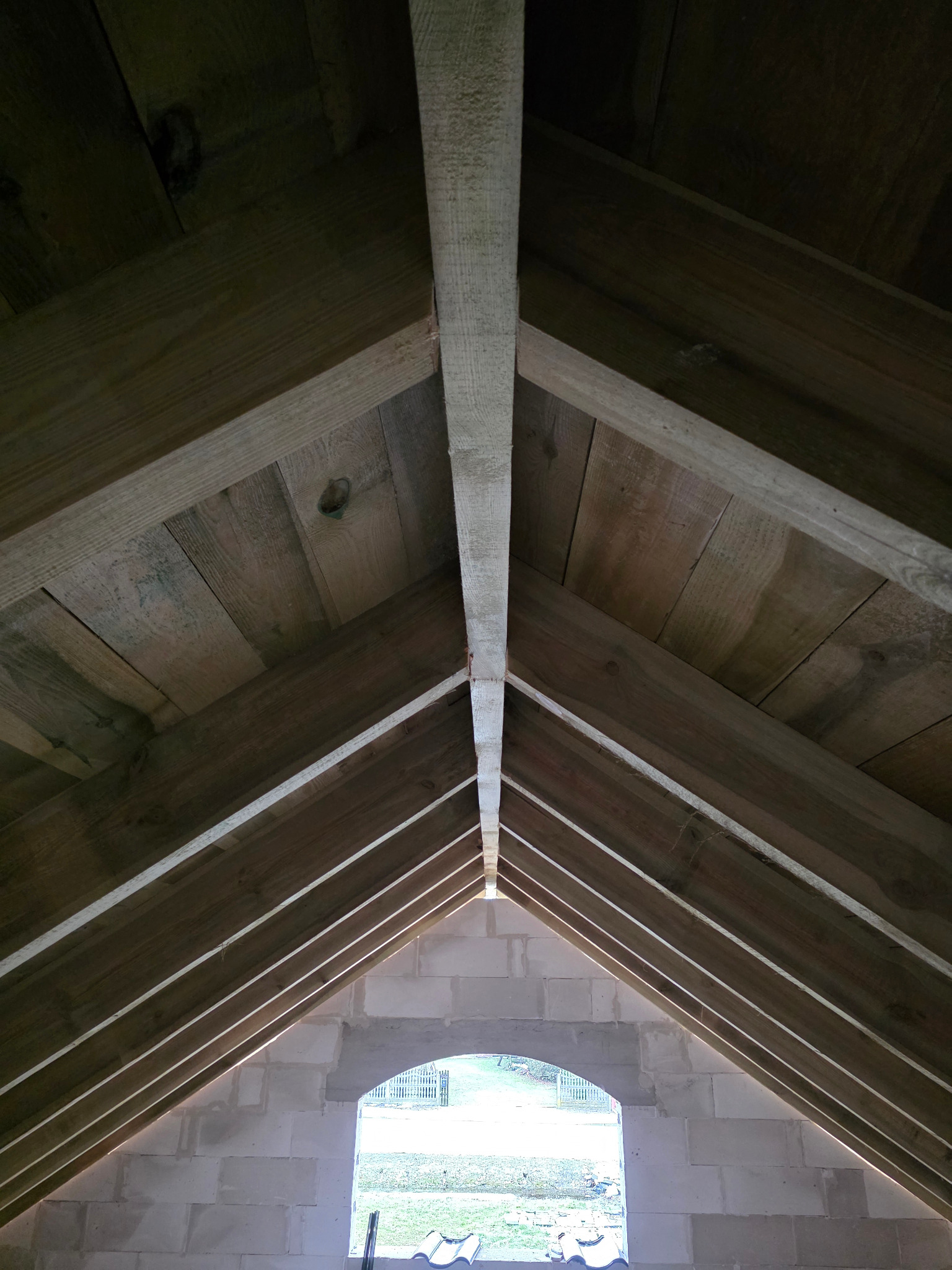 Wnętrze konstrukcji dachu z drewnianą więźbą i deskowaniem, widok od dołu, z oknem w tle. Surowy stan budowy, widoczne elementy konstrukcyjne.