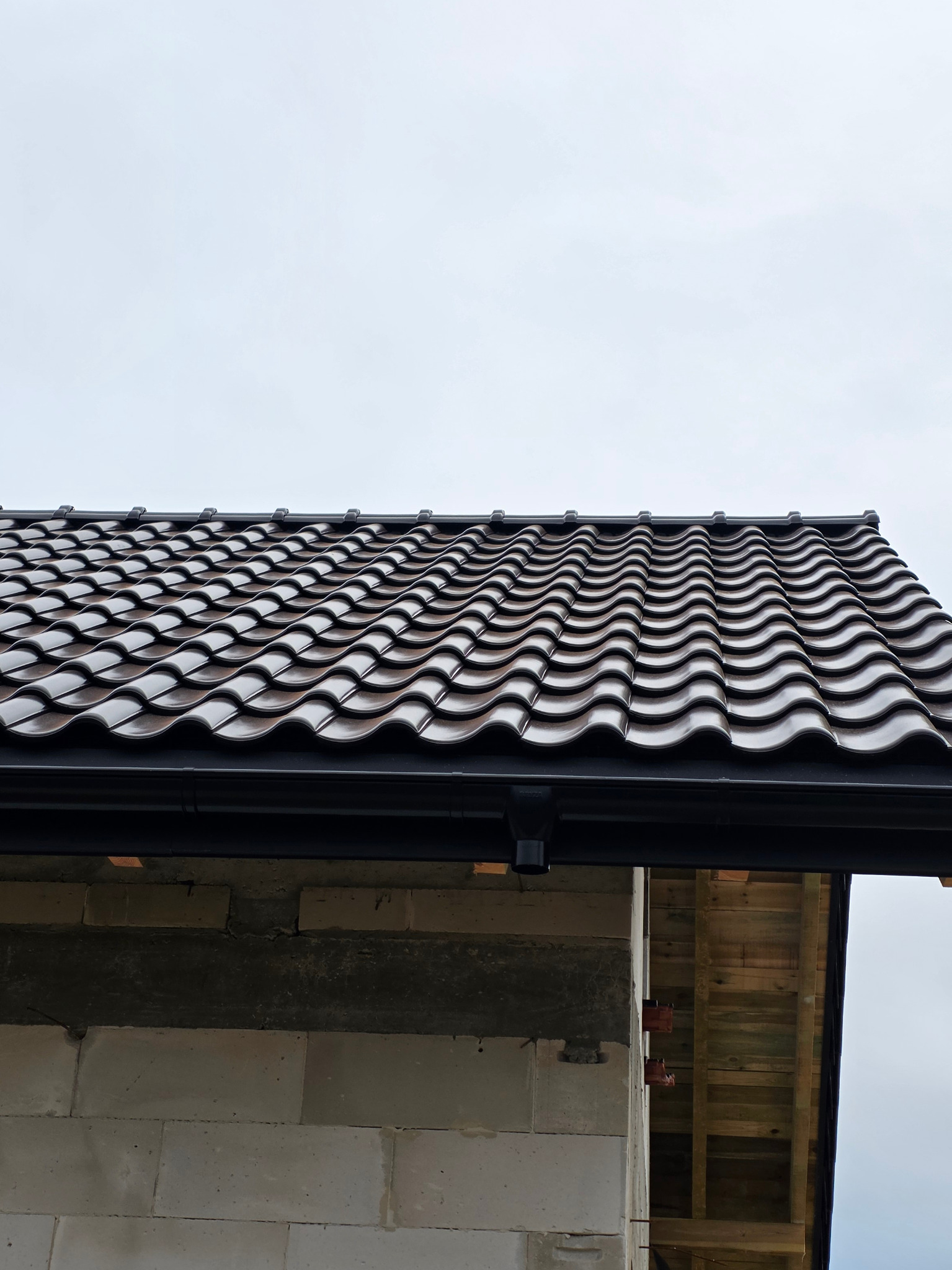 Dachówka ceramiczna Roben Milano w czarnym połysku, zamontowana na budynku z widoczną rynną. Ujęcie z dołu, podkreślające falisty wzór dachówki.