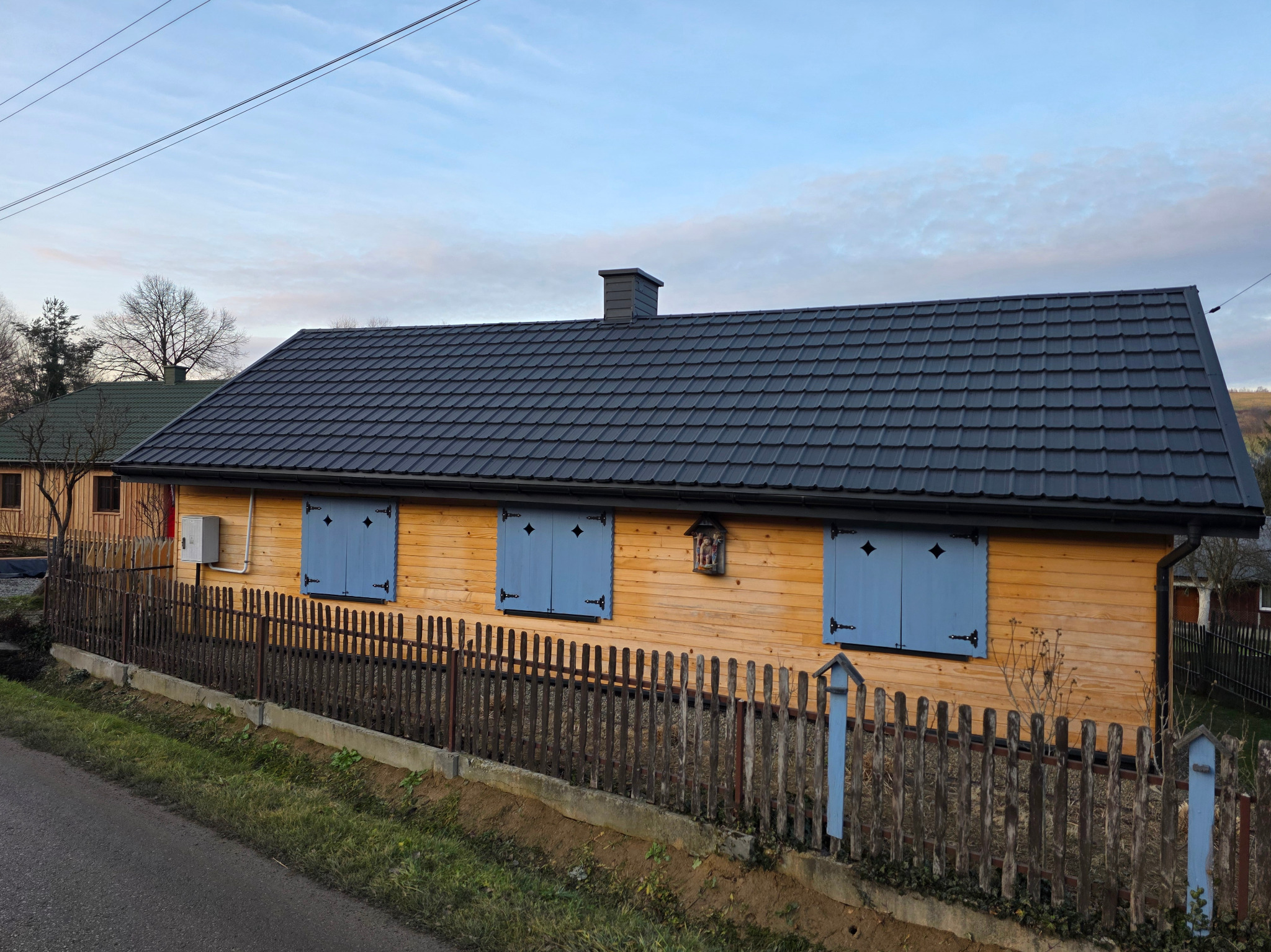 Dom z nowym, grafitowym dachem z blachy modułowej Finn 2.0, drewnianą elewacją i niebieskimi okiennicami. Widok z poziomu ulicy, w tle drzewa.