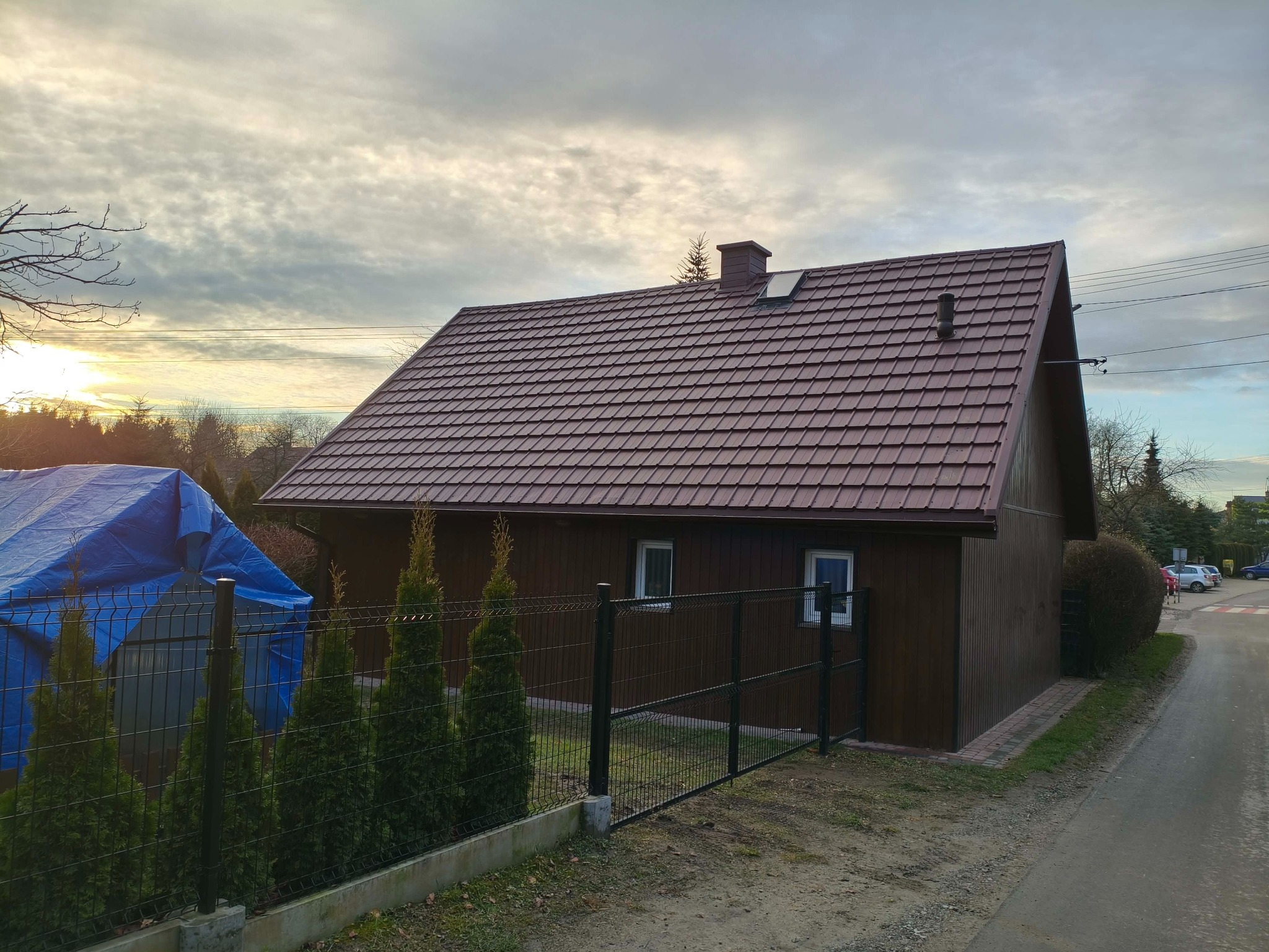 Dom z nowym, brązowym dachem z blachy KAN-POL Cortina Ultramat, otoczony ogrodzeniem i zielenią. W tle pochmurne niebo z przebijającym się słońcem.
