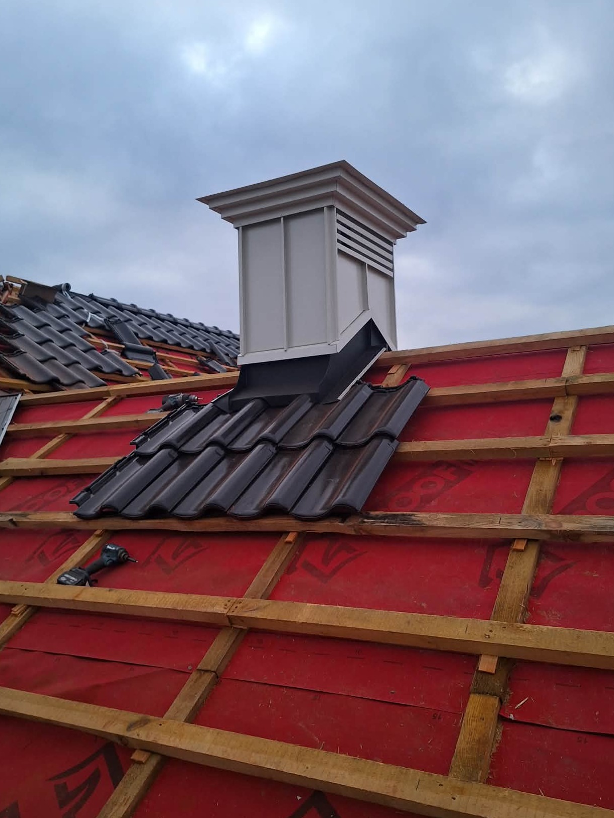 Okucie komina blachą w kolorze brudnej bieli na tle czarnej dachówki, widoczne elementy więźby i narzędzia. Prace dekarskie w trakcie realizacji.