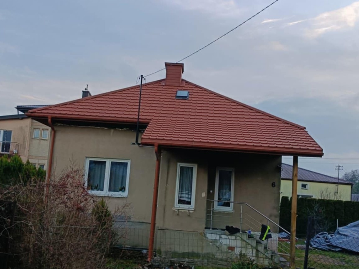 Dom z nowym, ceglastym dachem kopertowym FINN 2.0 BP2, z rynnami w kolorze dachu. Widoczne okna dachowe i komin. Tło: zabudowa jednorodzinna, pochmurne niebo.