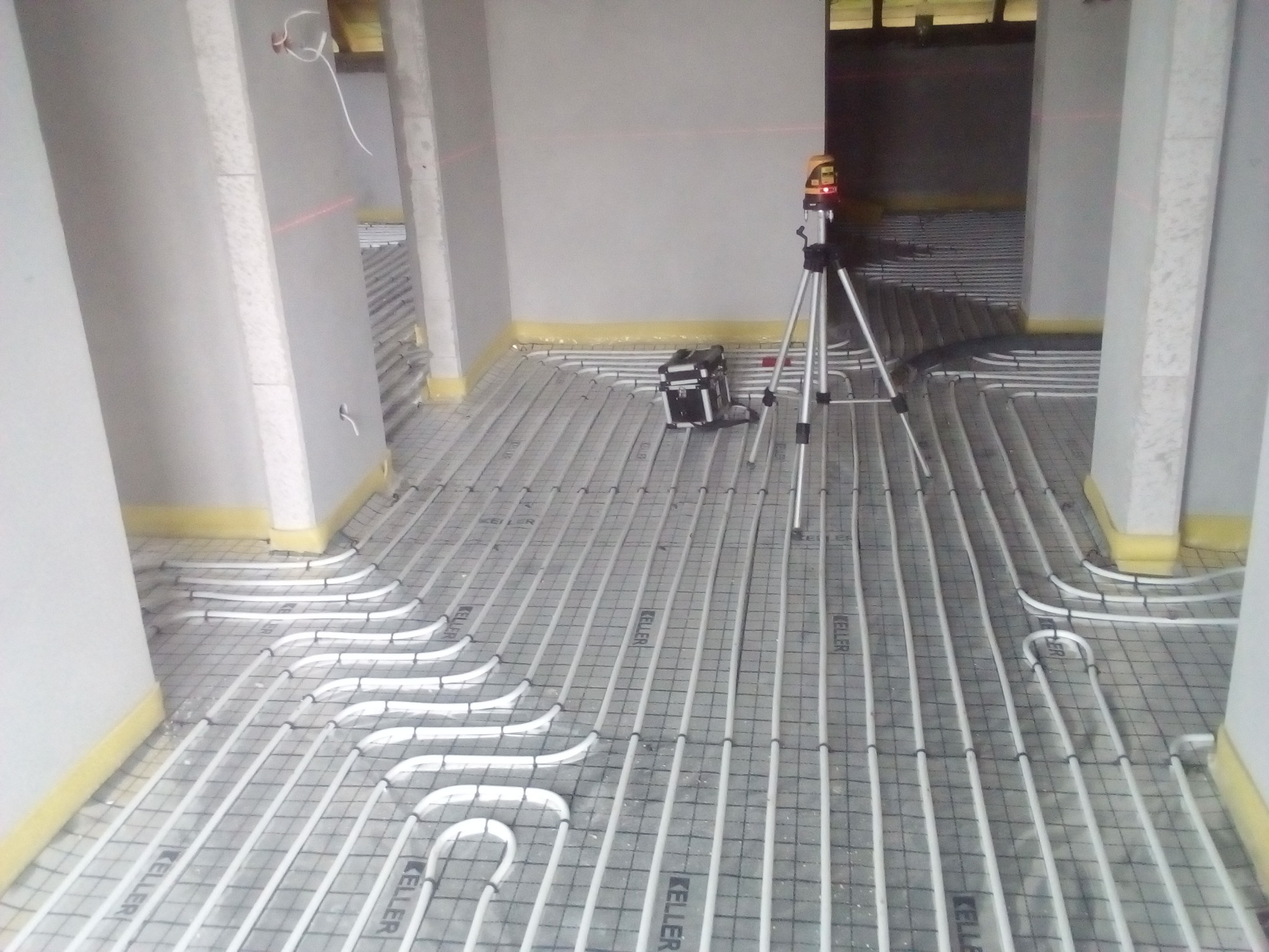 Rozłożone rury ogrzewania podłogowego na siatce, widoczny laser krzyżowy na statywie i skrzynka narzędziowa w surowym wnętrzu.