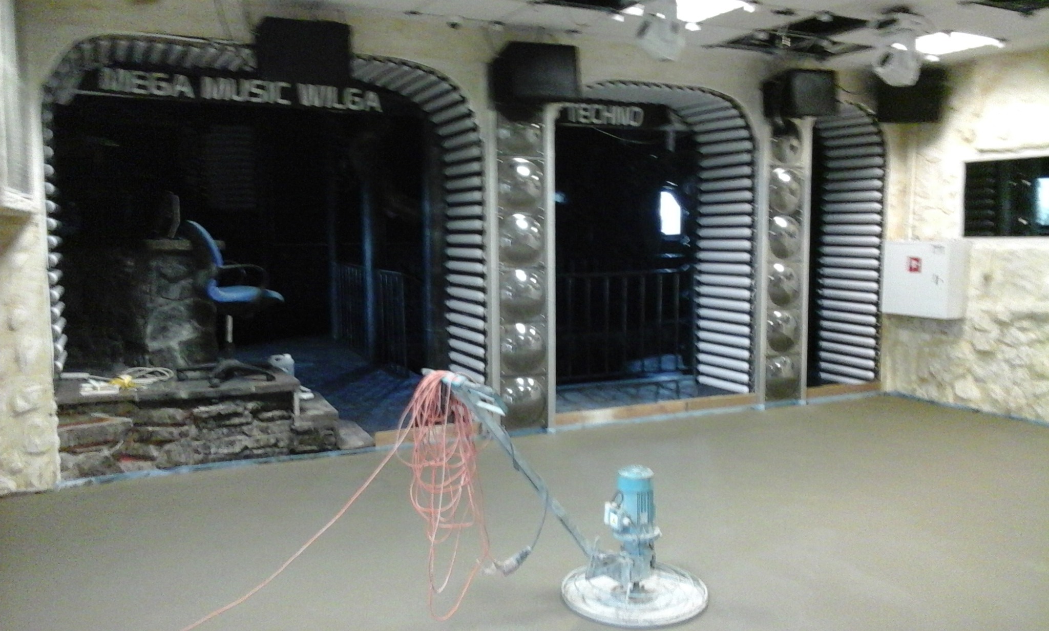 Szlifowanie wylewki w lokalu rozrywkowym, widoczna maszyna szlifująca z plątaniną kabli, w tle wejścia do pomieszczeń z dekoracjami.