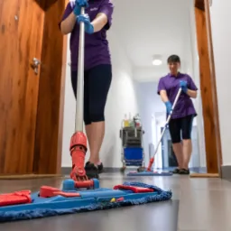 Dwie pracownice w fioletowych uniformach myją podłogę mopami w korytarzu biurowym; w tle wózek ze środkami czystości i drewniane drzwi.