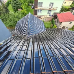 Błyszczący, ciemny dach z dachówki ceramicznej, widok z góry na fragment dachu z kominem i otoczeniem domów jednorodzinnych.