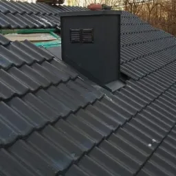 Czarny komin wentylacyjny z kratkami na dachu z ciemnej dachówki ceramicznej, widoczne fragmenty konstrukcji dachu i narzędzia.