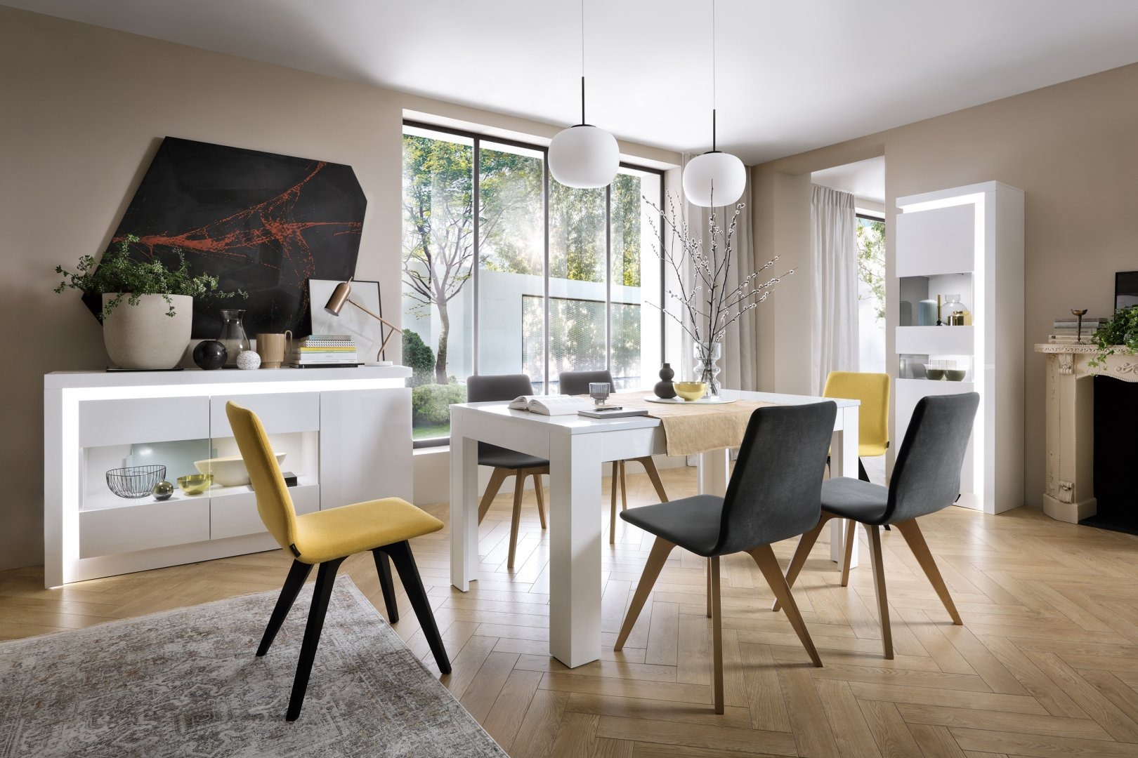 Nowoczesna jadalnia z białym stołem, szarymi i żółtymi krzesłami, oświetleniem kulistym i parkietem w jodełkę, z widokiem na ogród przez duże okno.