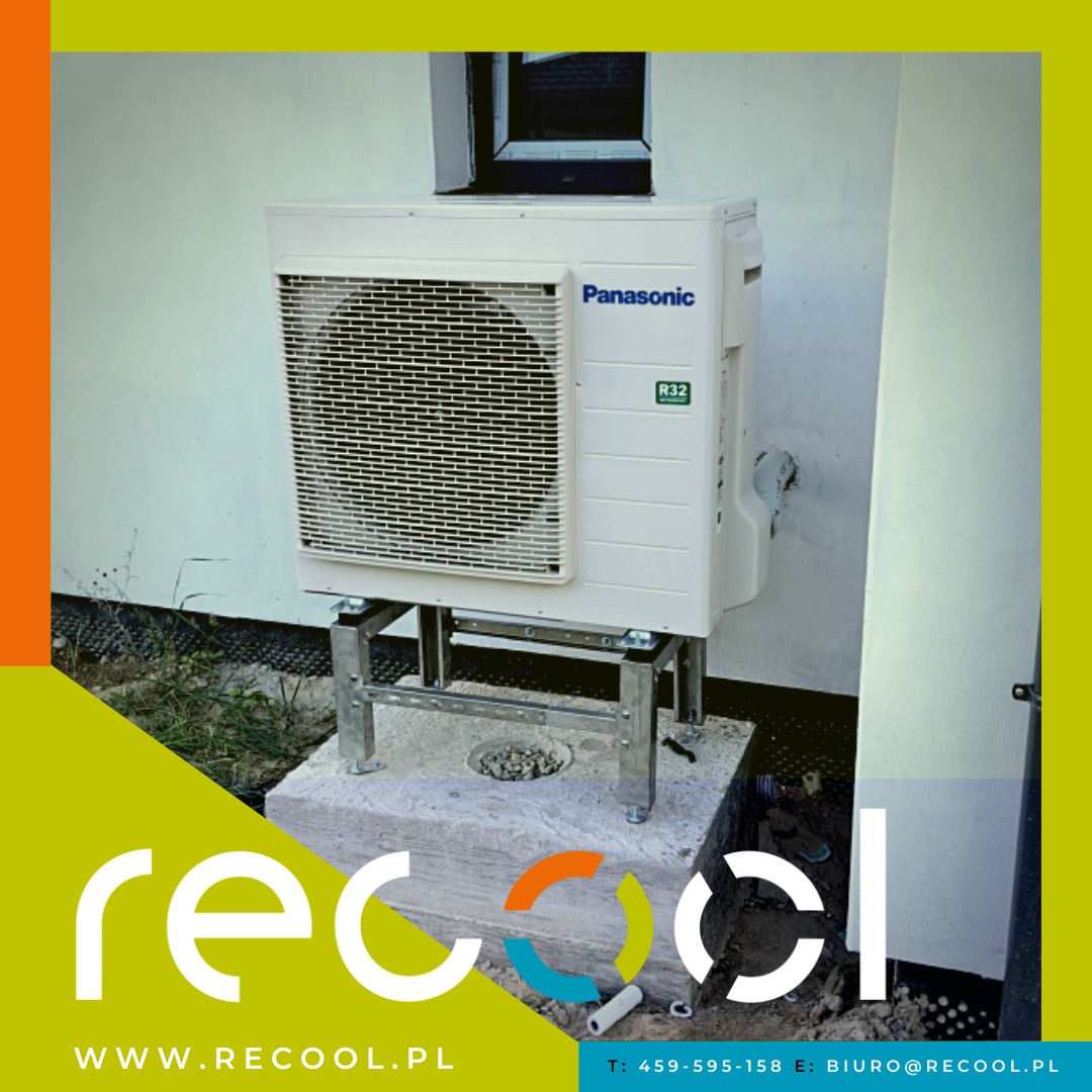Zewnętrzna jednostka klimatyzacyjna Panasonic z czynnikiem chłodniczym R32, zamontowana na metalowej konstrukcji na betonowej podstawie, przy białej ścianie budynku z małym oknem.