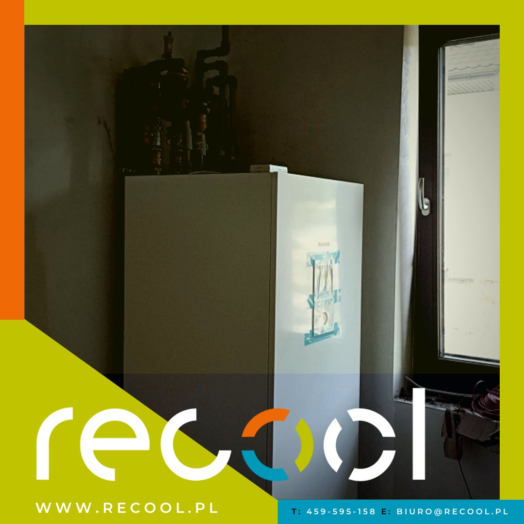 Biały bojler wiszący na ścianie, z widocznymi rurami instalacji i fragmentem okna po prawej stronie, zabezpieczony taśmą montażową, z logo firmy na pierwszym planie.