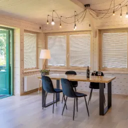 Jasne wnętrze z drewnianymi panelami, trzema oknami z białymi żaluzjami poziomymi, stołem z butelką wina i kieliszkami, oświetleniem sufitowym z żarówkami na kablach oraz otwartymi drzwiami...